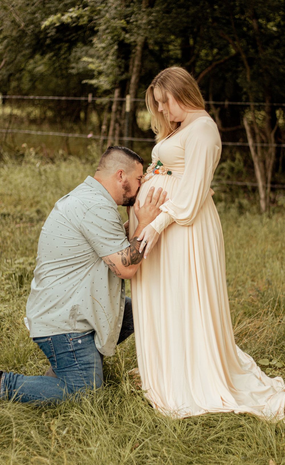 A tender maternity moment captured outdoors as a couple embraces in a grassy field, wearing cream and grey clothing.
