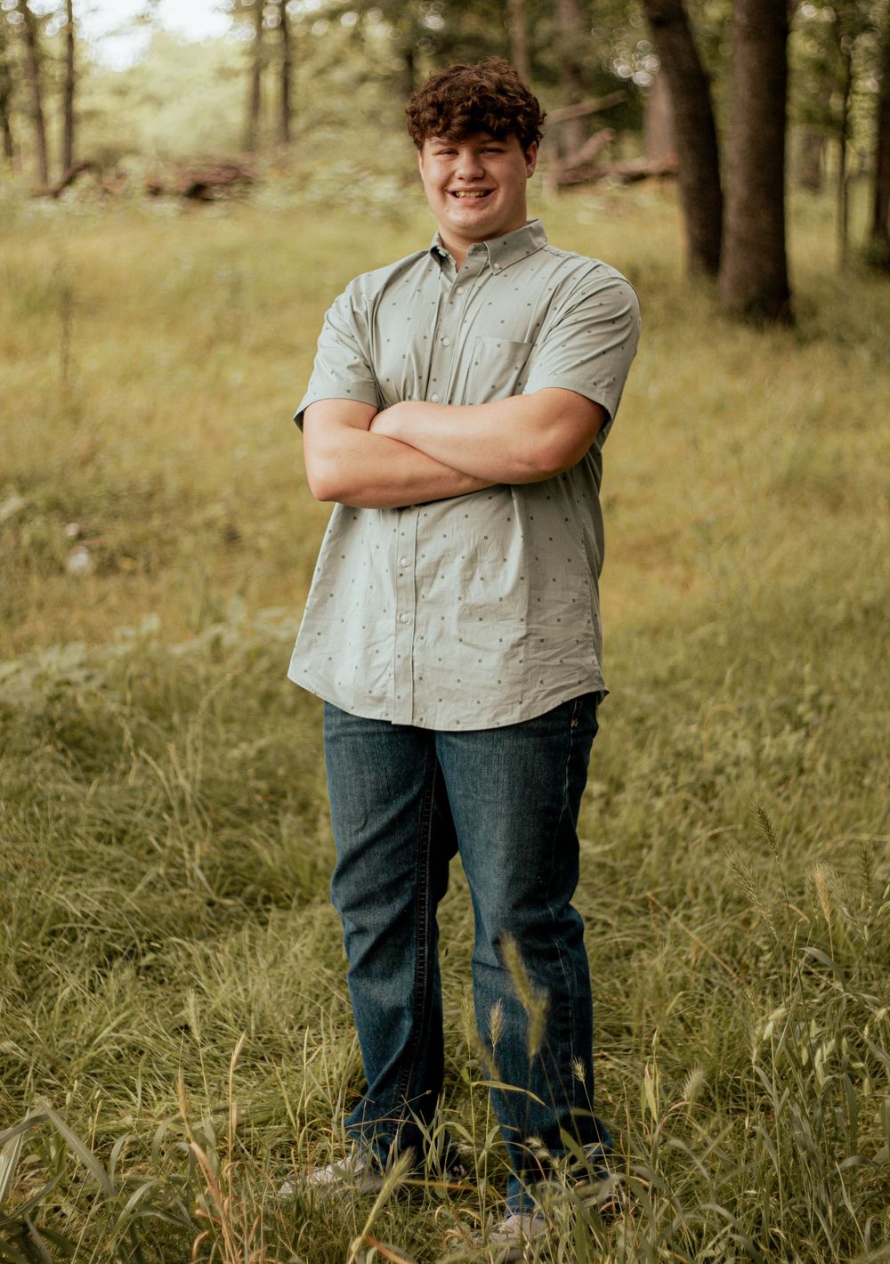Someone wearing a light gray button-down shirt and jeans stands with crossed arms in a wooded outdoor setting.