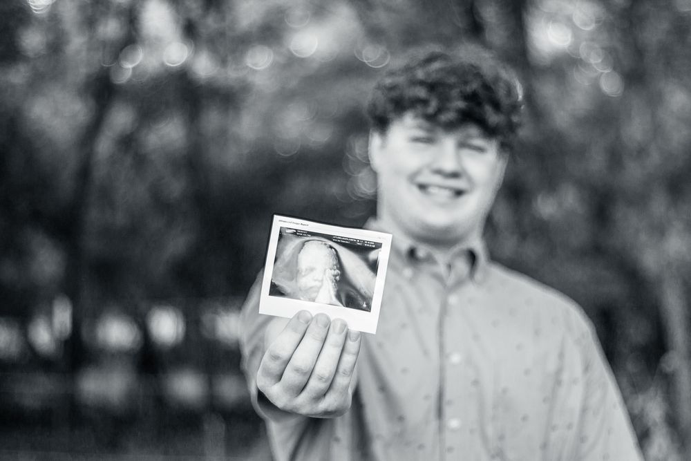 A person stands outdoors holding up a small instant photograph in a black and white shot.