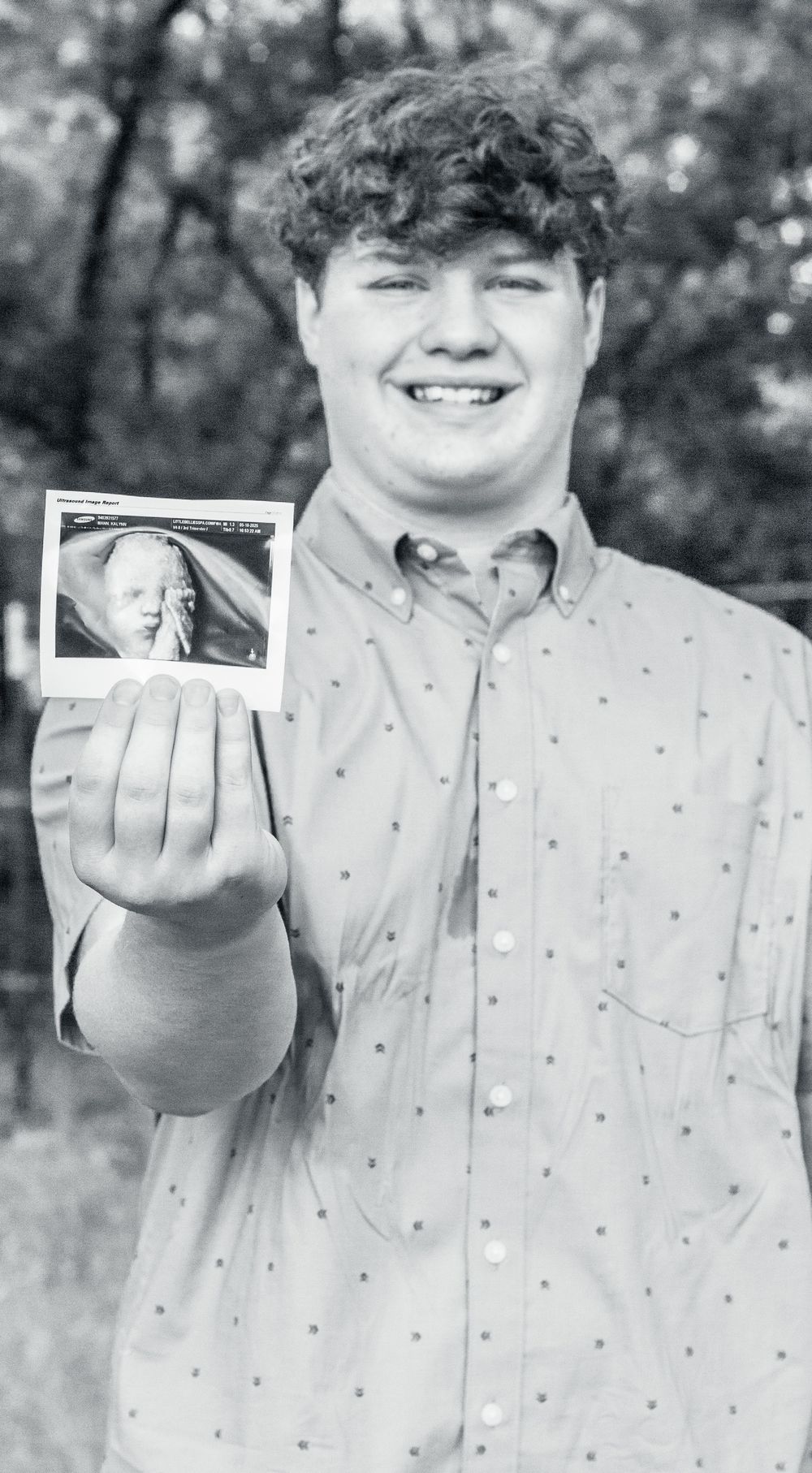 A person in a button-down shirt holds up a small polaroid photograph while standing outdoors.