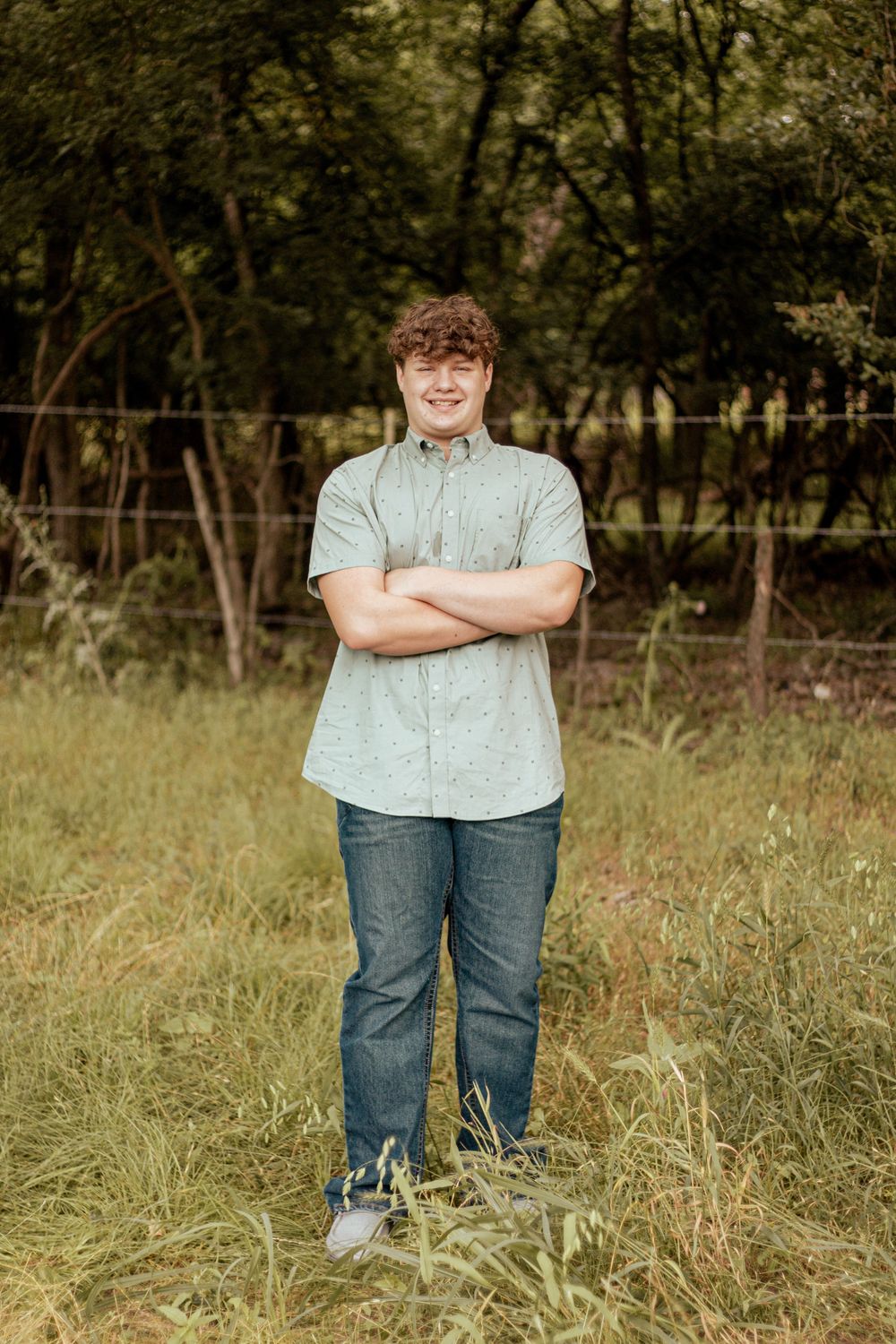 A person stands casually in light shirt and jeans in a sunlit grassy field with trees in the background.