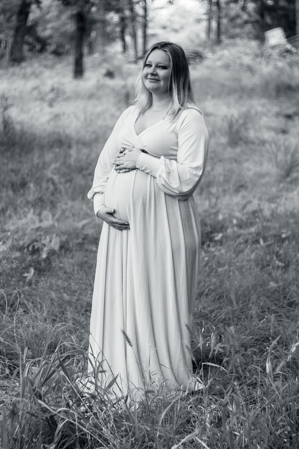 A black and white maternity photo shows an expectant mother in a flowing white dress standing in a forest setting.