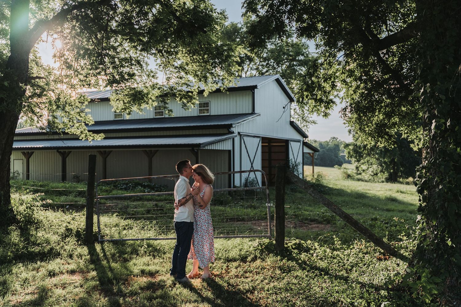 A couple embraces and laughs together on a sunny day in front of a rural farmhouse with green trees in the background.