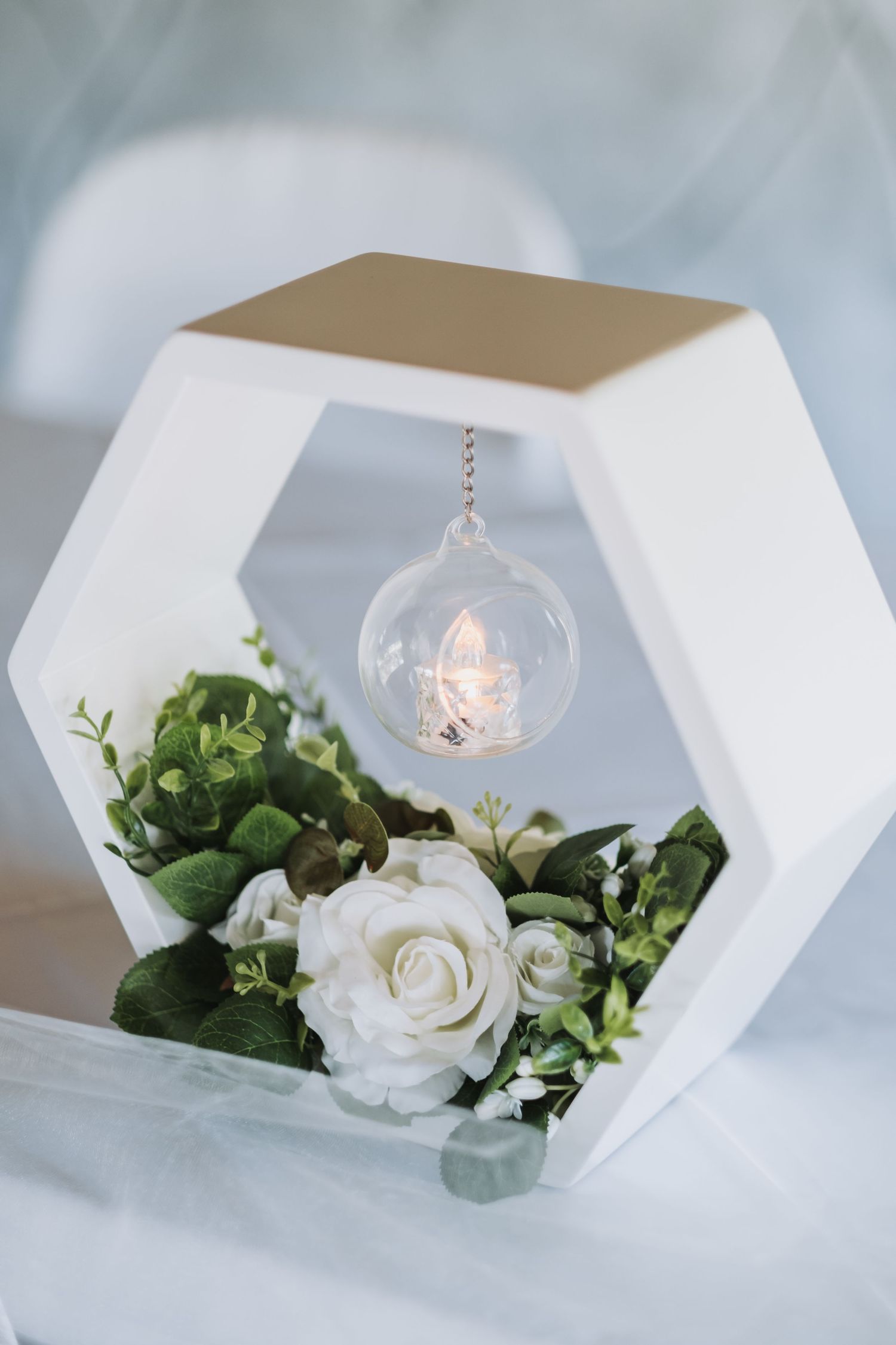 White hexagonal wedding centerpiece with white roses, greenery, and a hanging glass globe candle holder.