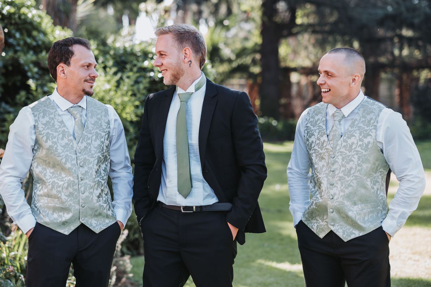Three groomsmen laughing together outdoors wearing black suits with silver paisley vests and sage green ties.