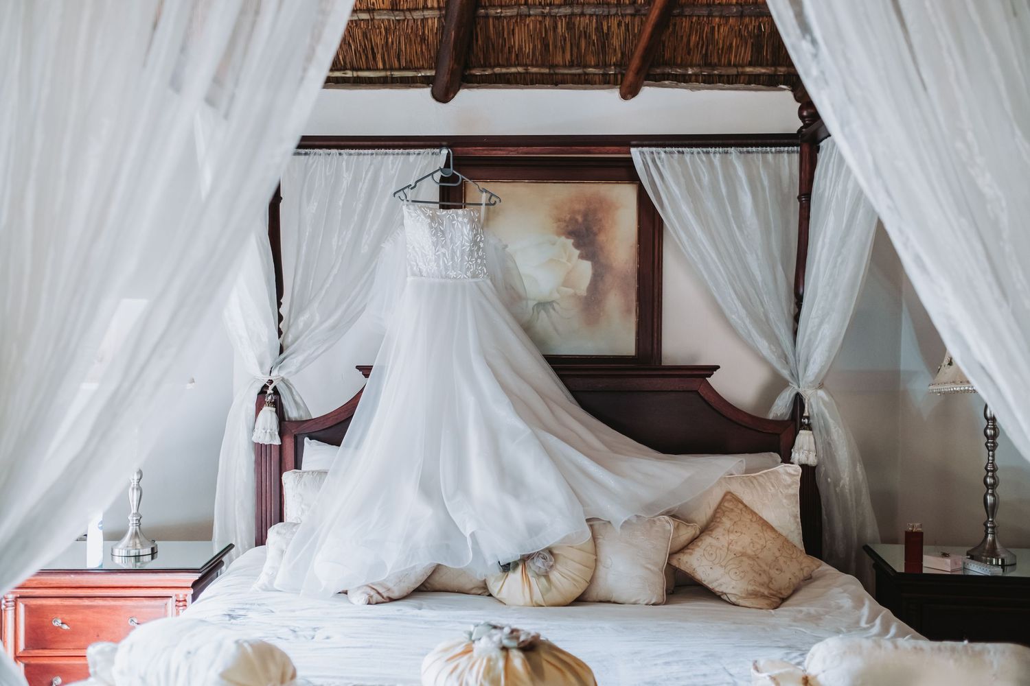 Elegant white wedding gown hanging on a four-poster bed in a rustic thatched-roof bridal suite.