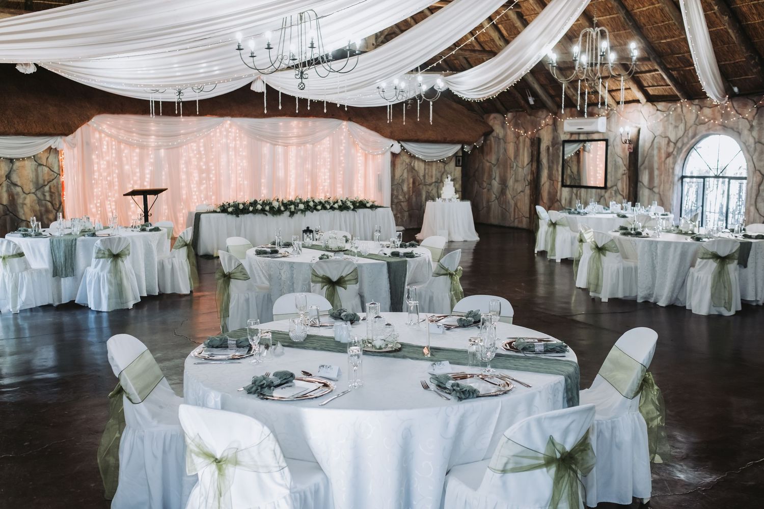 Elegant wedding reception hall with white draped ceiling, round tables, sage chair sashes, and chandelier.