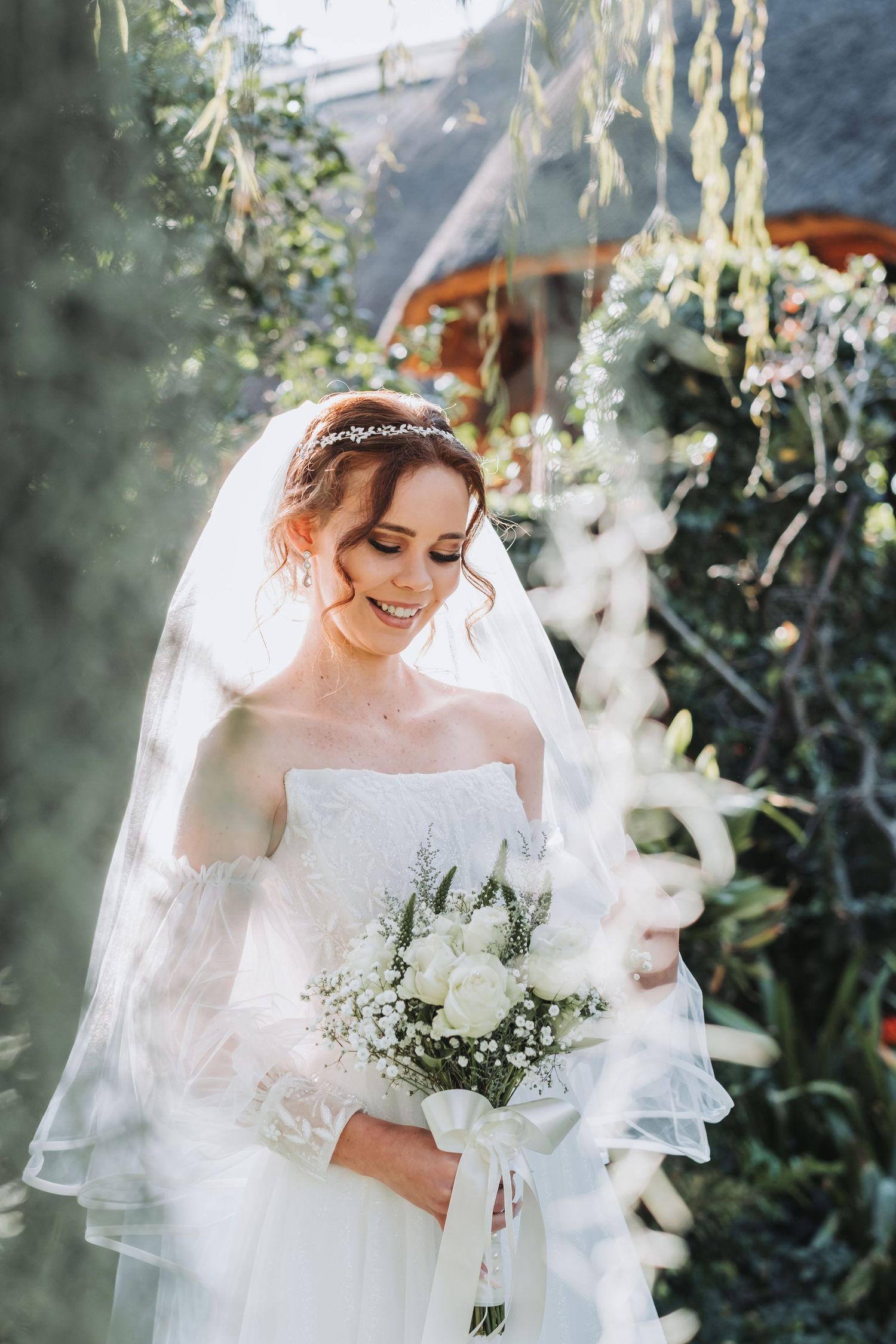 Smiling bride in white off-shoulder gown and veil holds white bouquet outdoors in sunlit garden.