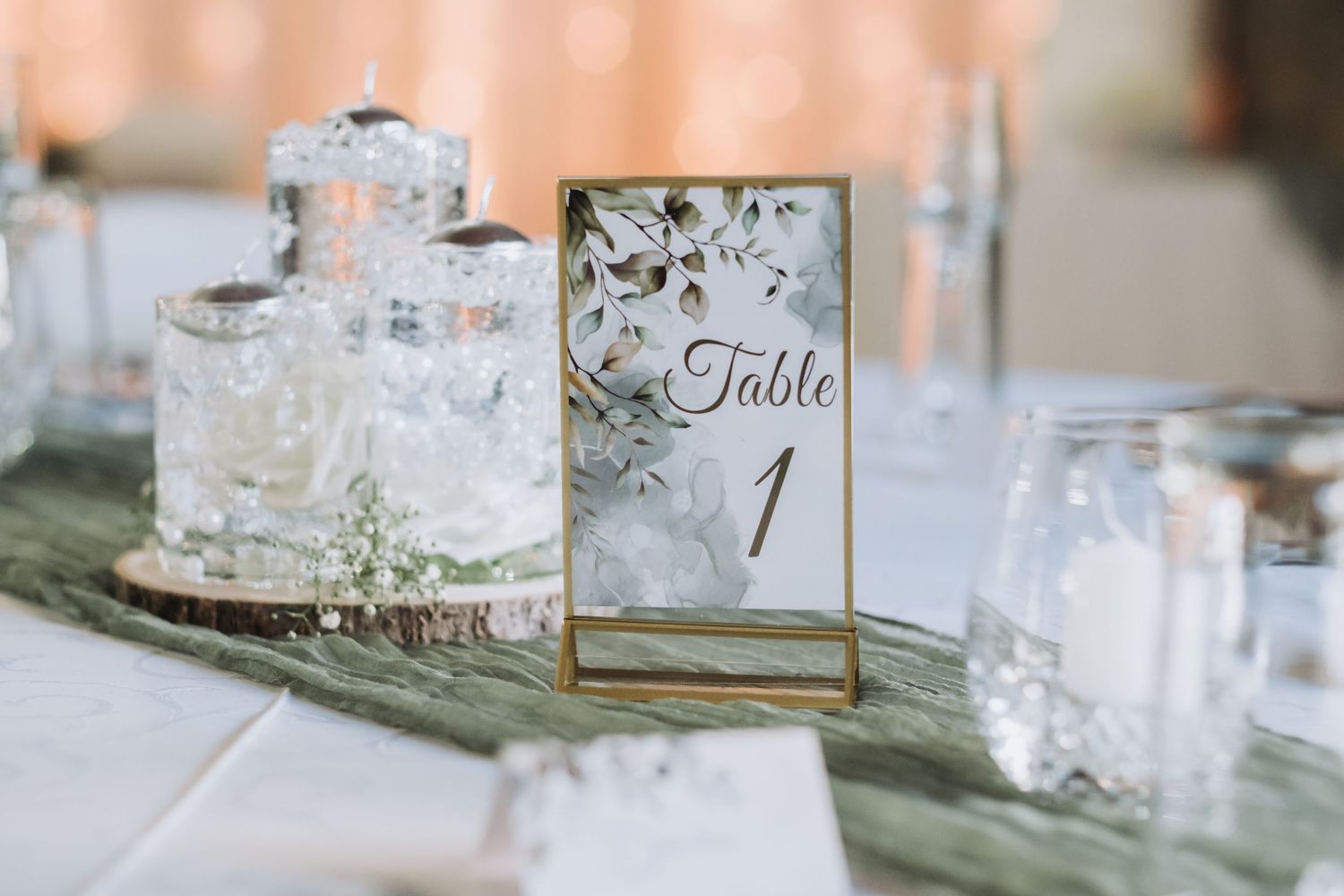 Gold-framed Table 1 sign with floral design displayed on a sage green runner with crystal decorations.