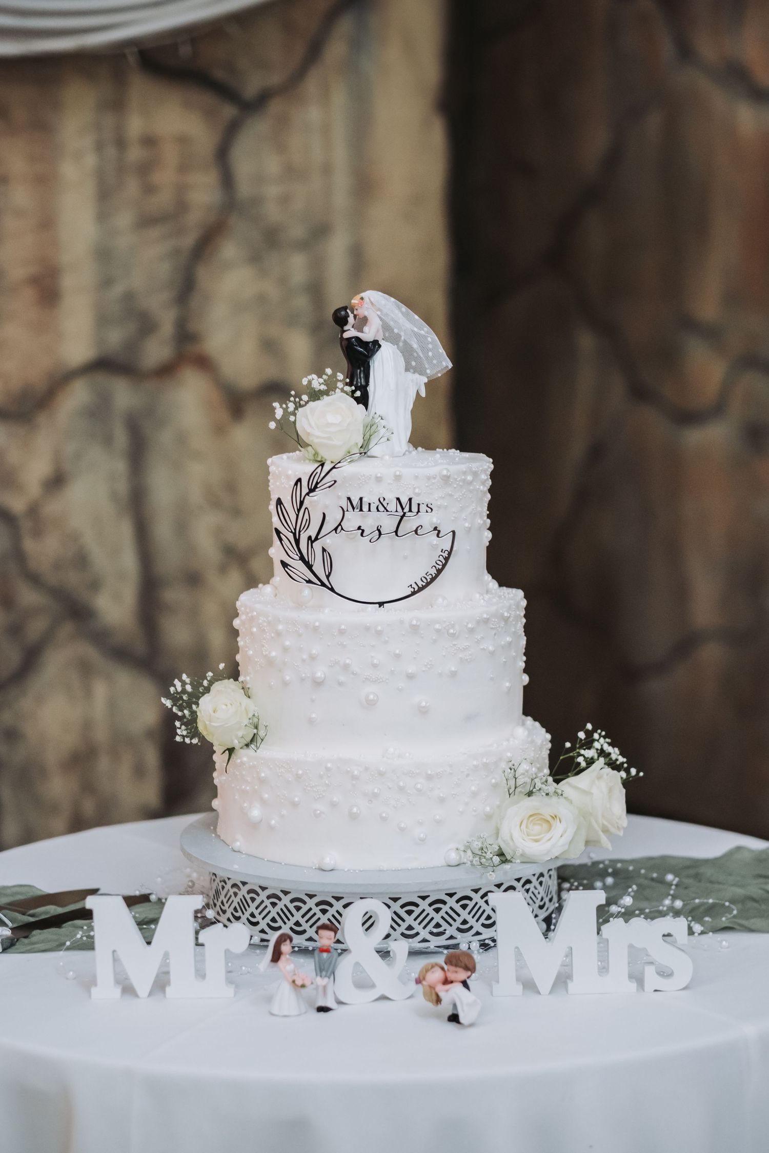 Three-tier white wedding cake with pearl details, floral topper, and Mr & Mrs sign on a white stand.