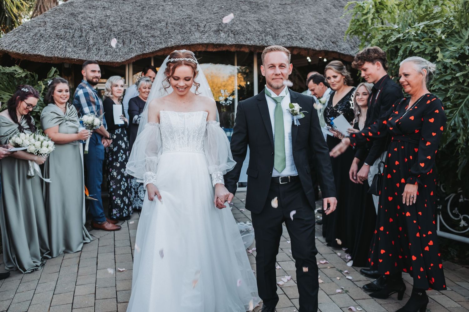 Newlyweds walk hand in hand through confetti shower with wedding party guests lining the path.