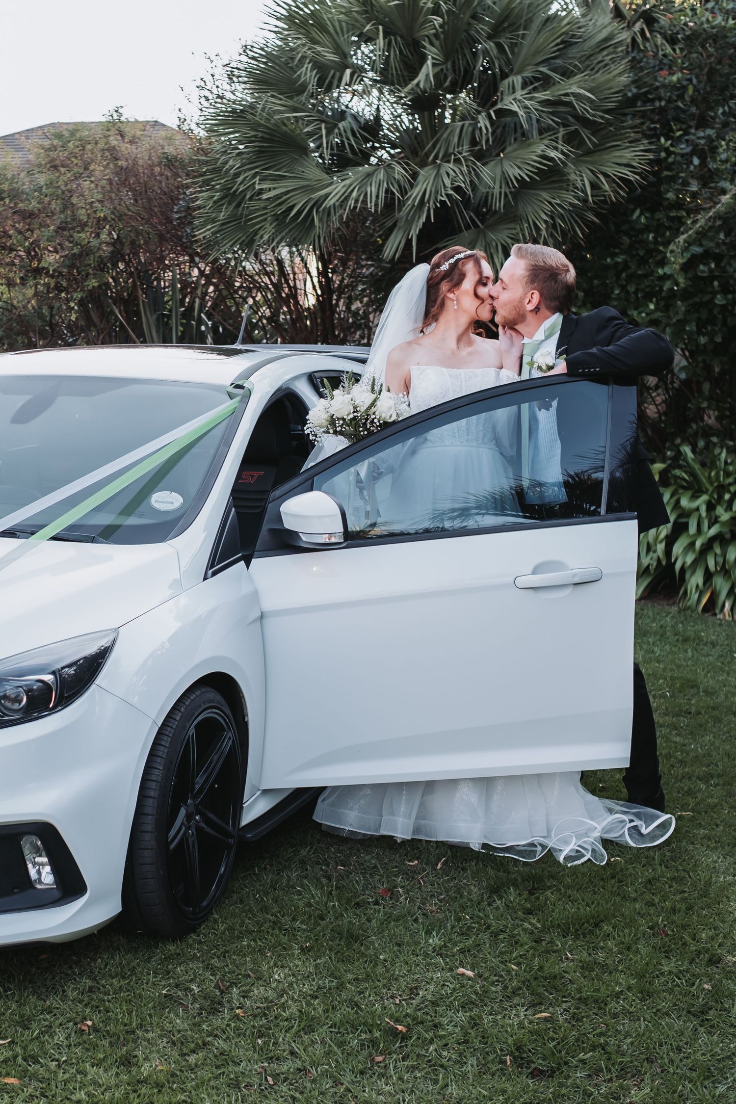 Bride and groom share a kiss beside a decorated white car in a lush garden setting.