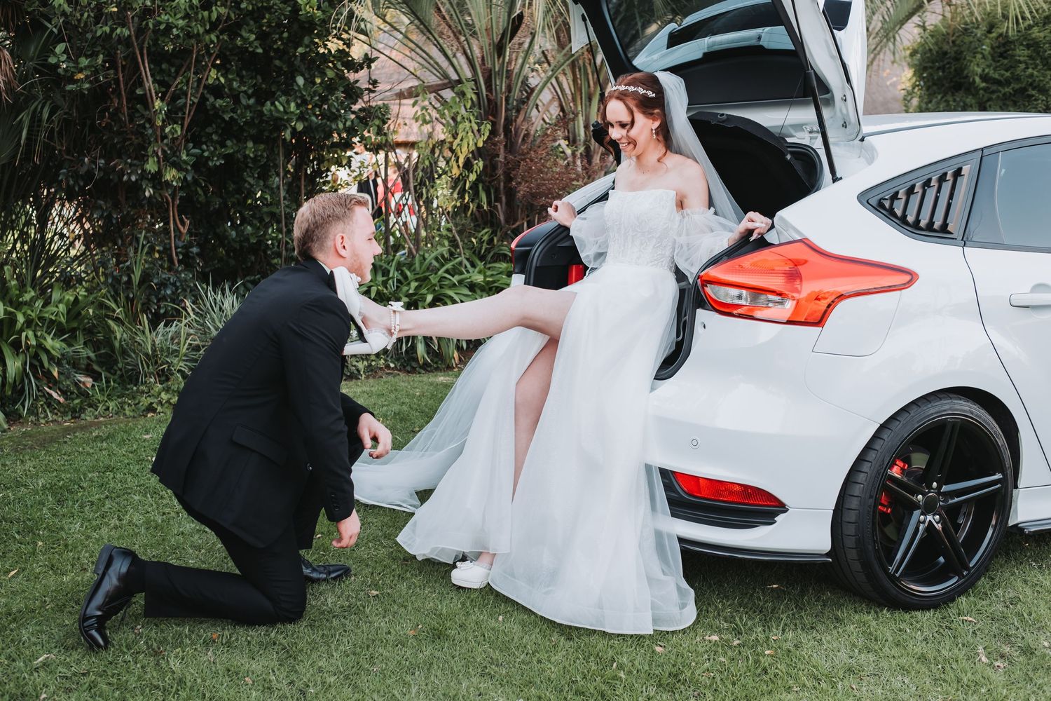 Groom kneels playfully beside bride sitting on white car trunk in outdoor wedding photo.