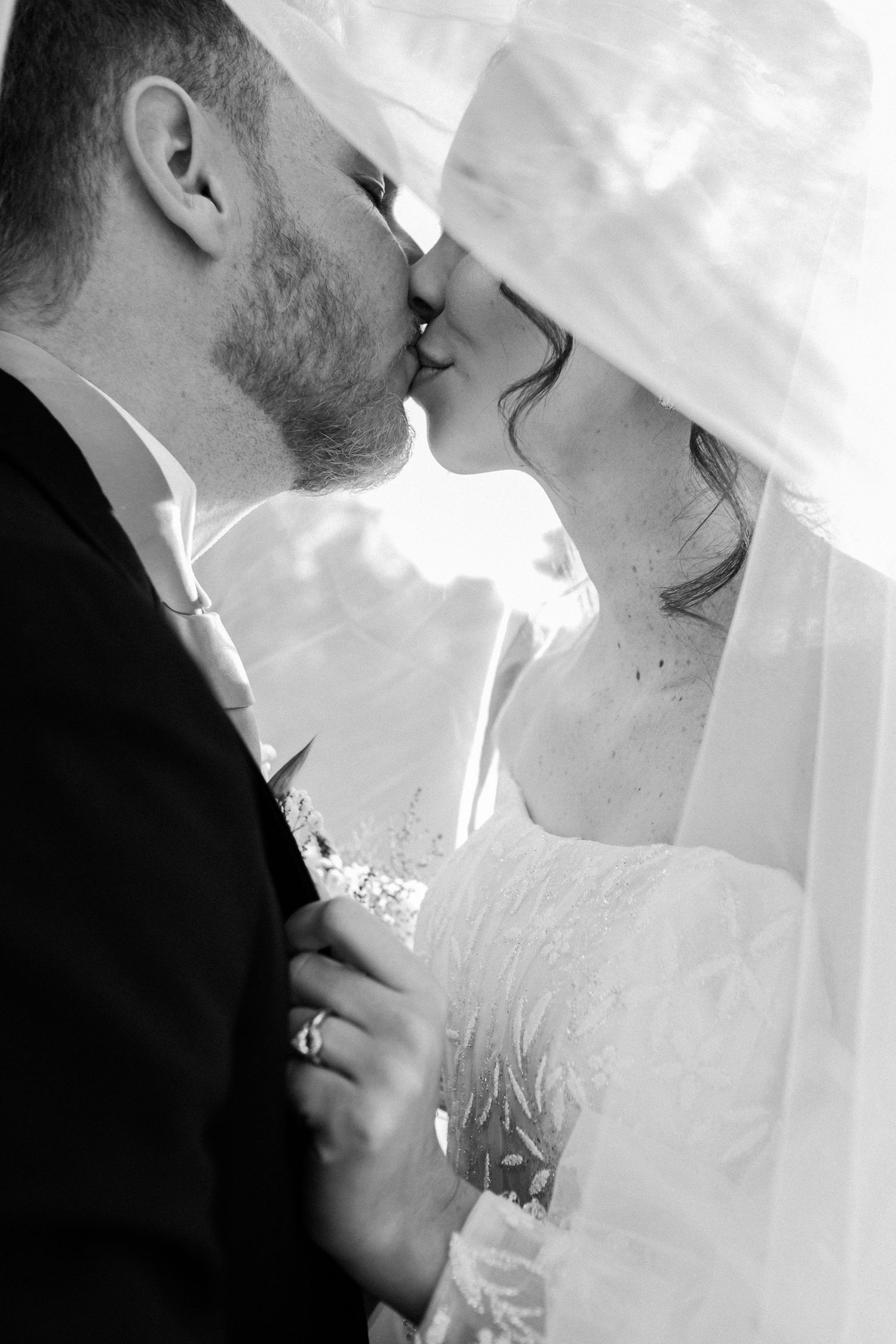 Black and white romantic photo of bride and groom kissing under flowing wedding veil.