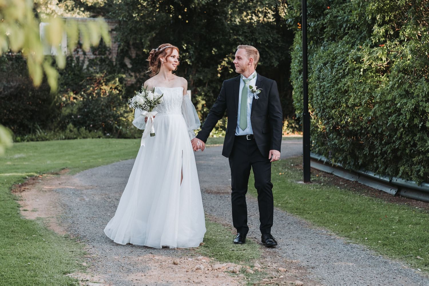 Bride and groom walk hand in hand along a garden path in soft golden hour light.