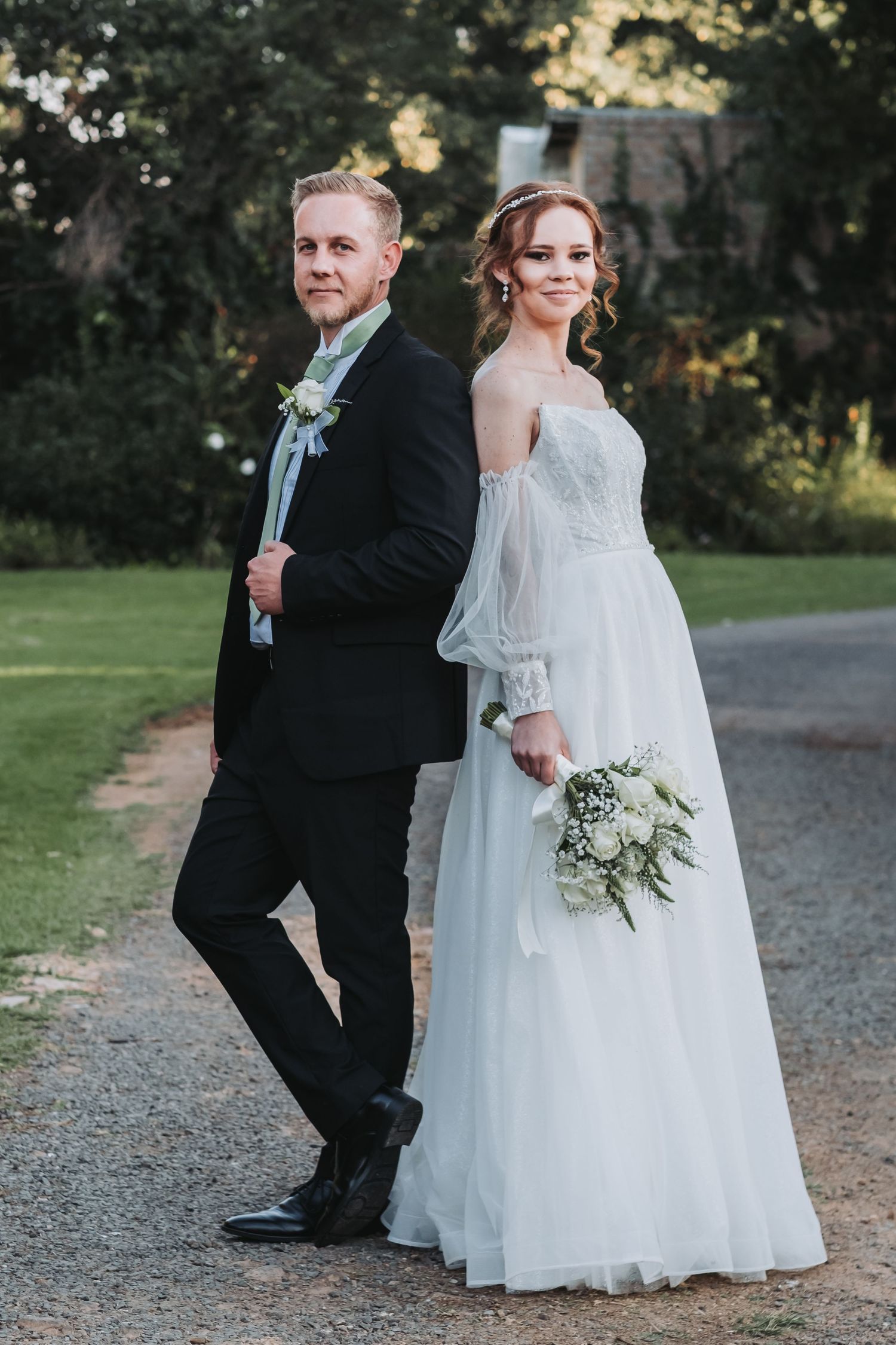 Bride holding white bouquet and groom in black suit pose back-to-back on gravel path.
