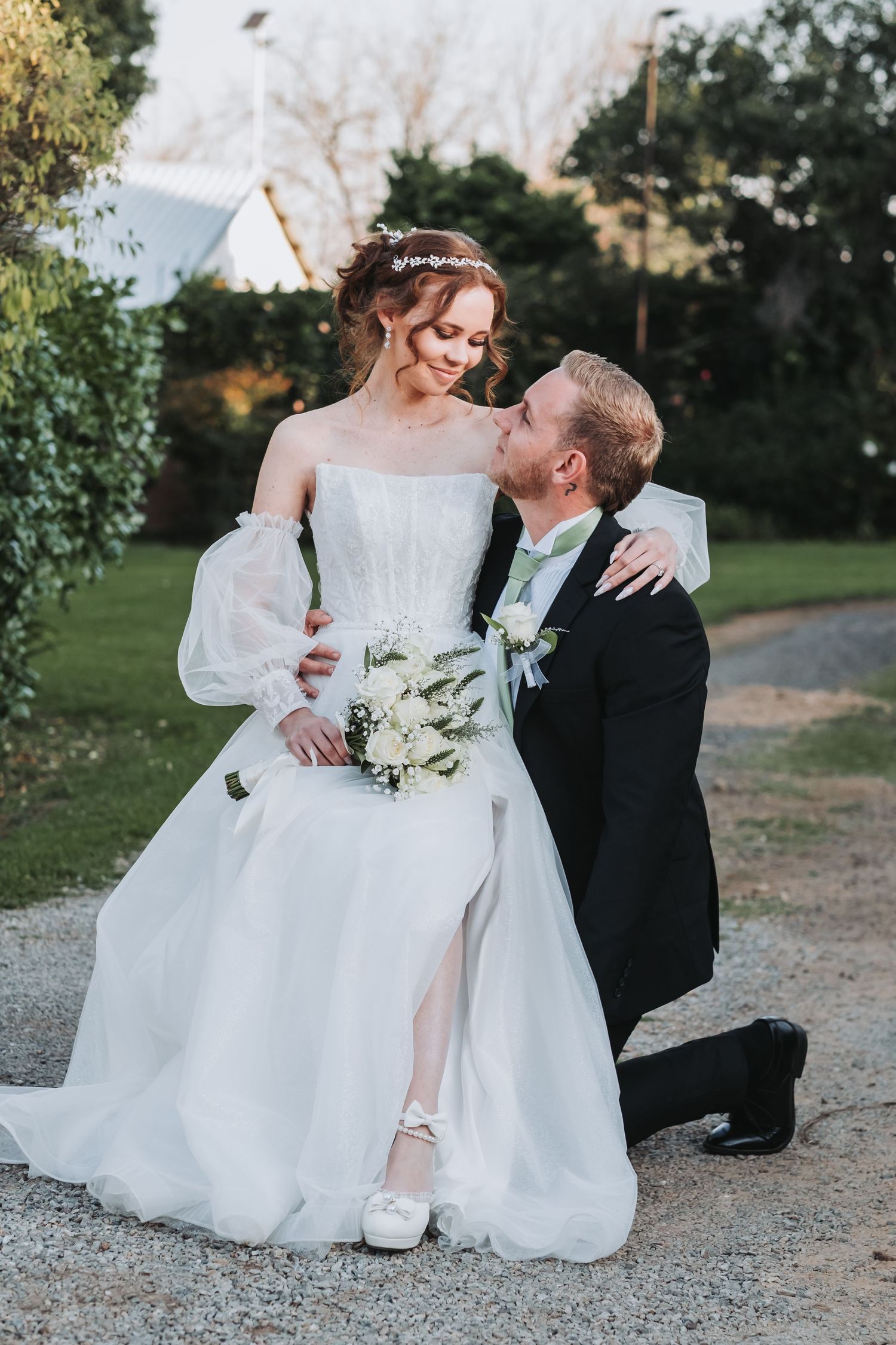 Groom kneels on gravel path gazing up at smiling bride holding white floral bouquet outdoors.