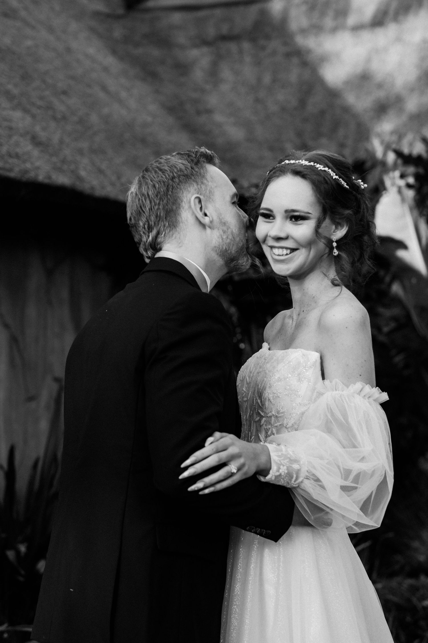 Black and white photo of groom whispering to laughing bride near rustic thatched building.
