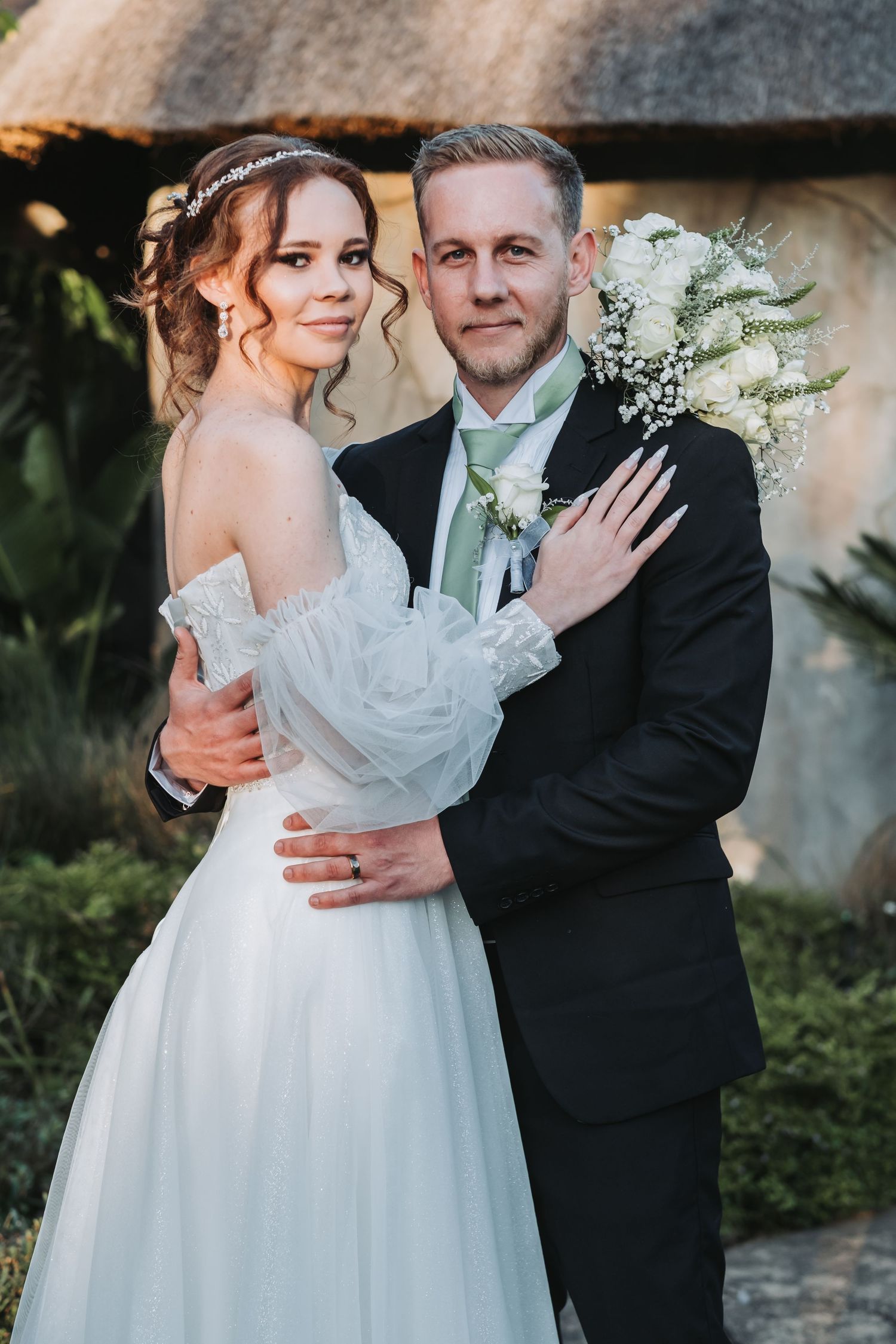 Bride and groom embrace warmly in front of thatched building surrounded by lush greenery.