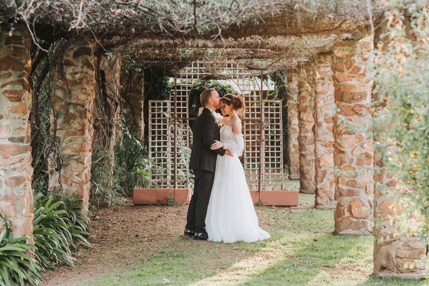 Bride and groom embracing romantically under a rustic stone pergola surrounded by lush greenery at their wedding.