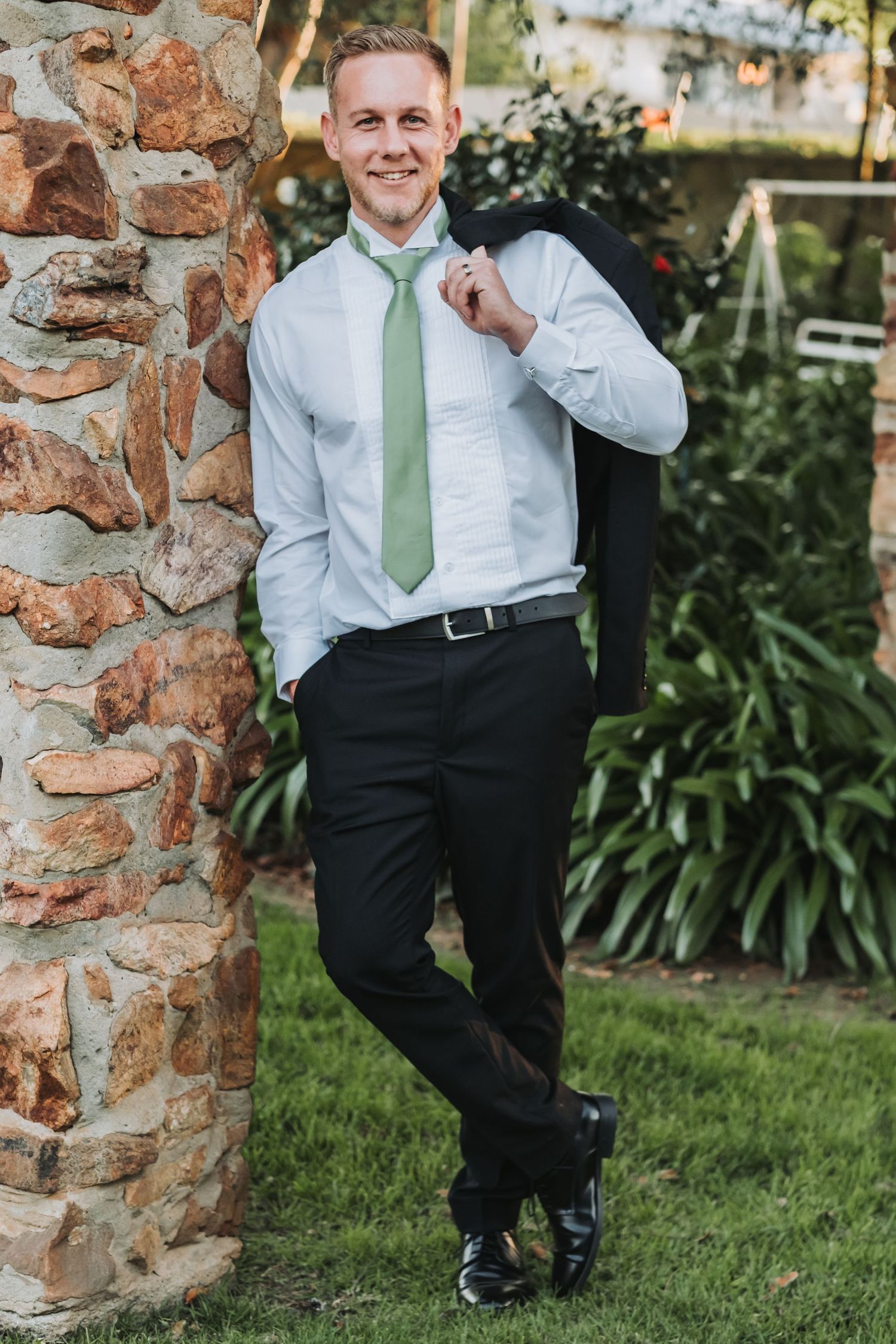 Handsome groom in white shirt and green tie holding suit jacket over shoulder, leaning against stone pillar outdoors.