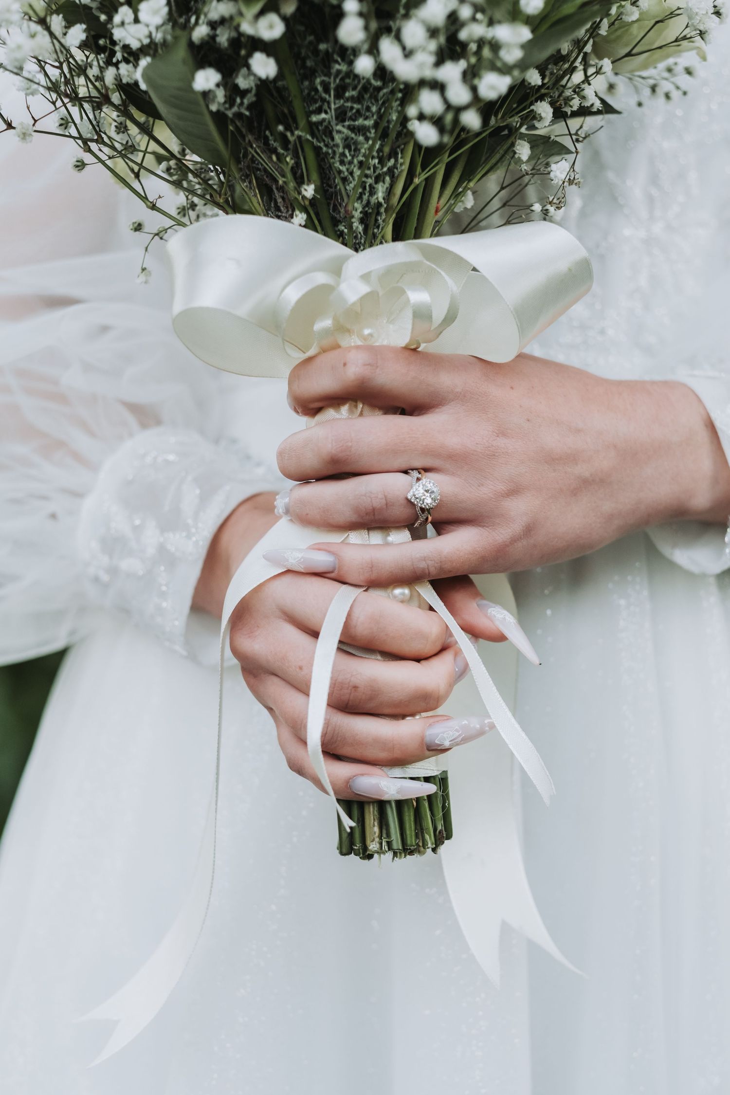 Close-up of bride holding white floral bouquet with ribbon, showcasing diamond engagement ring and manicured nails.