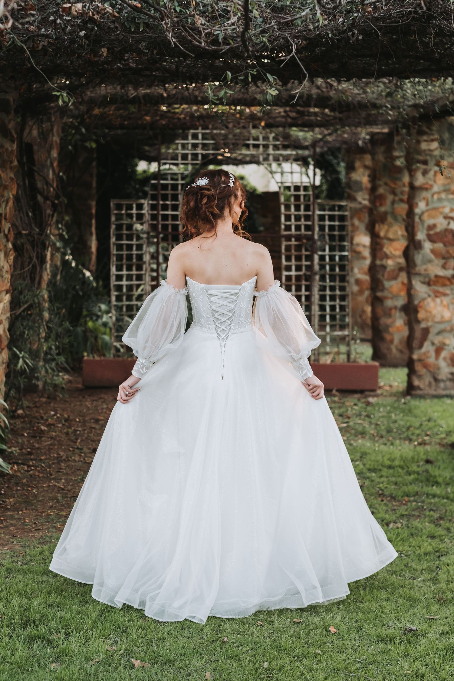 Back view of bride in stunning white corset ballgown with sheer sleeves standing under rustic garden pergola.