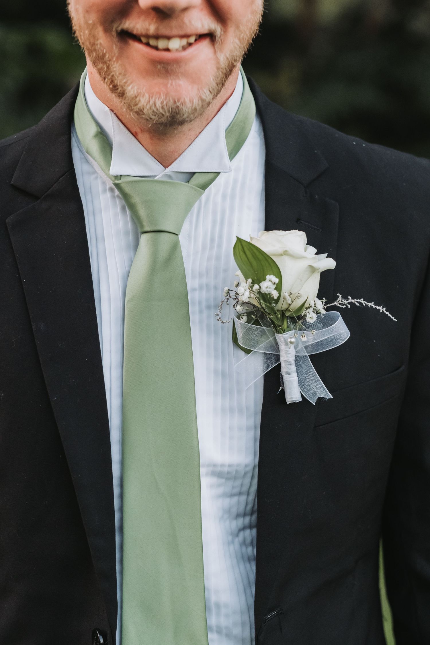 Close-up of groom in black suit with green tie and white rose boutonniere smiling at wedding reception.