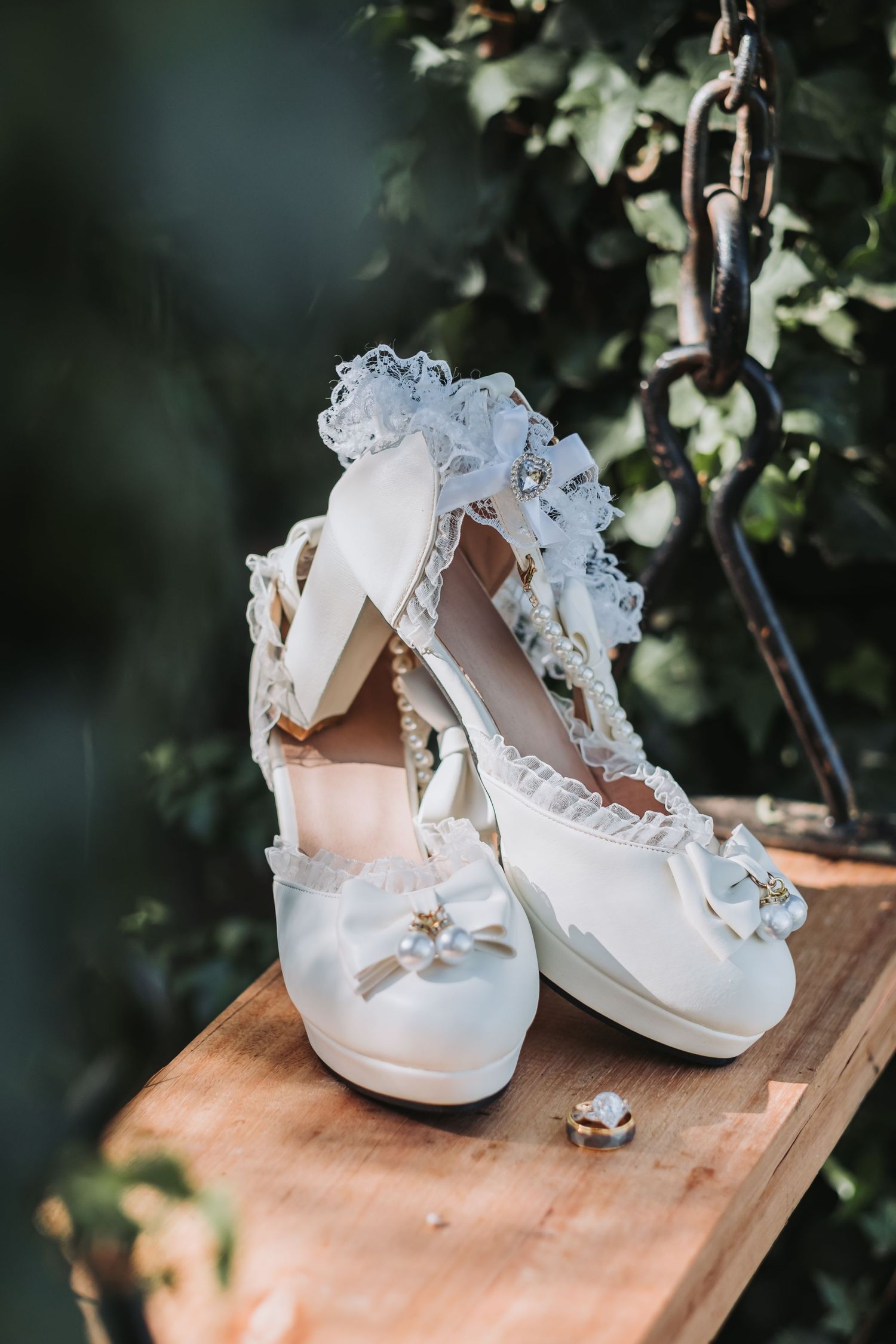 Elegant white bridal heels with lace details and bow embellishments displayed on a wooden surface outdoors.