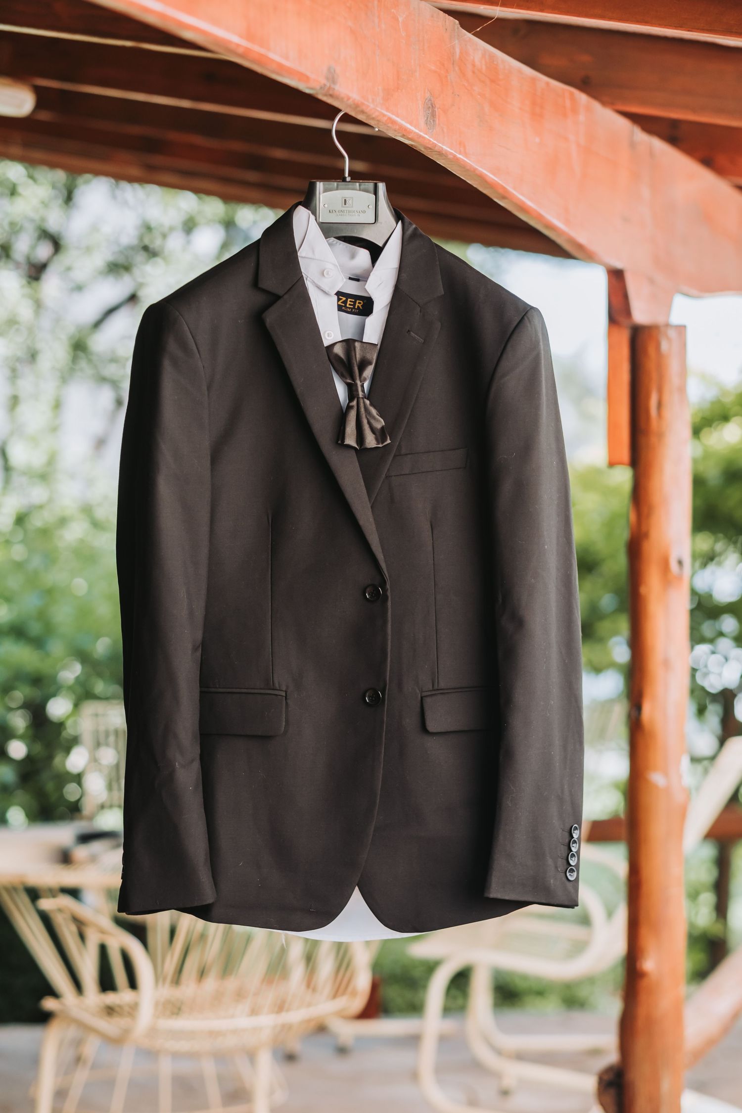 Dark charcoal groom's suit jacket hanging on a wooden beam with a patterned tie outdoors.