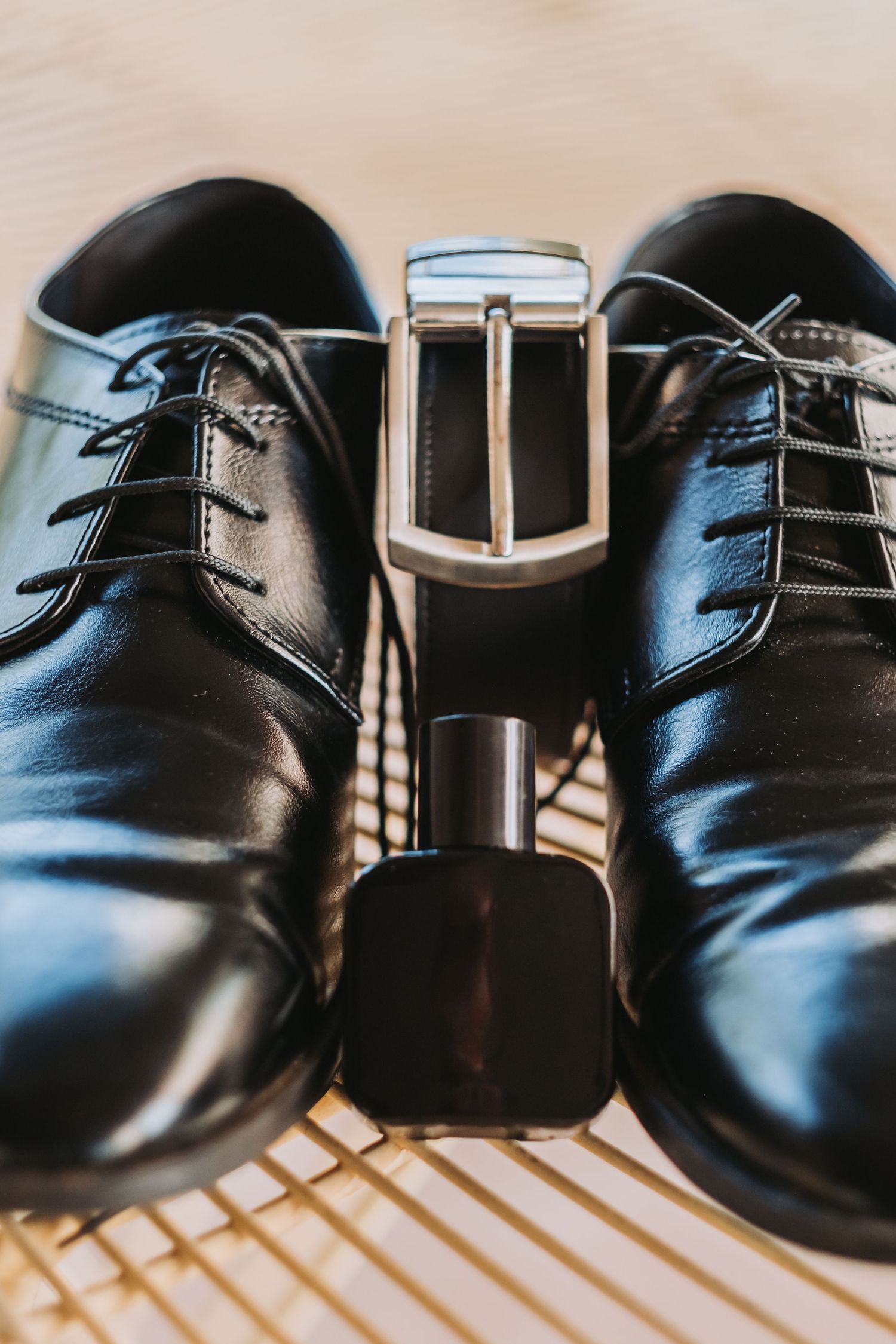 Polished black leather dress shoes with a matching belt and cologne bottle arranged on a surface.