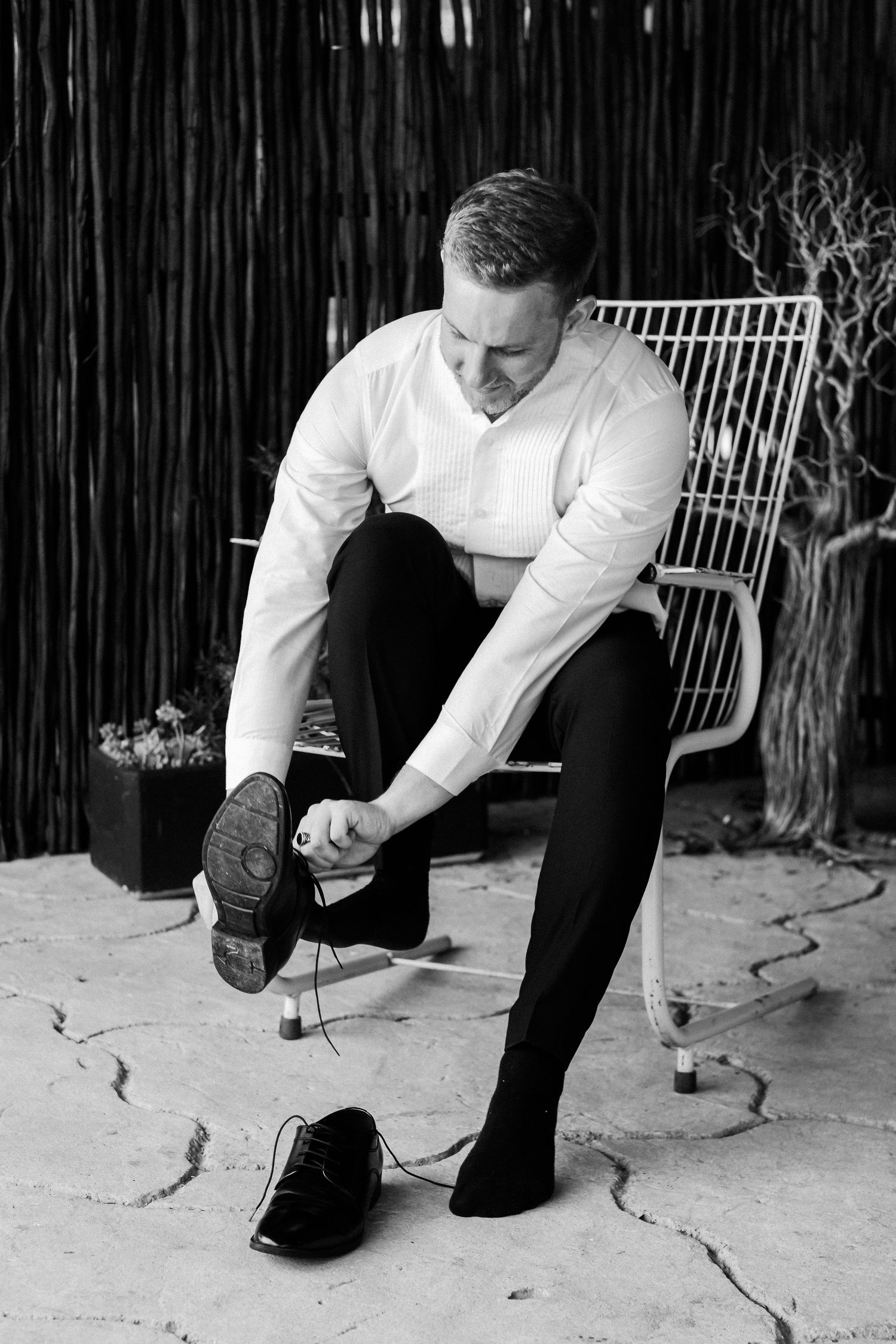 Black and white photo of a groom putting on his dress shoes while seated on a chair.