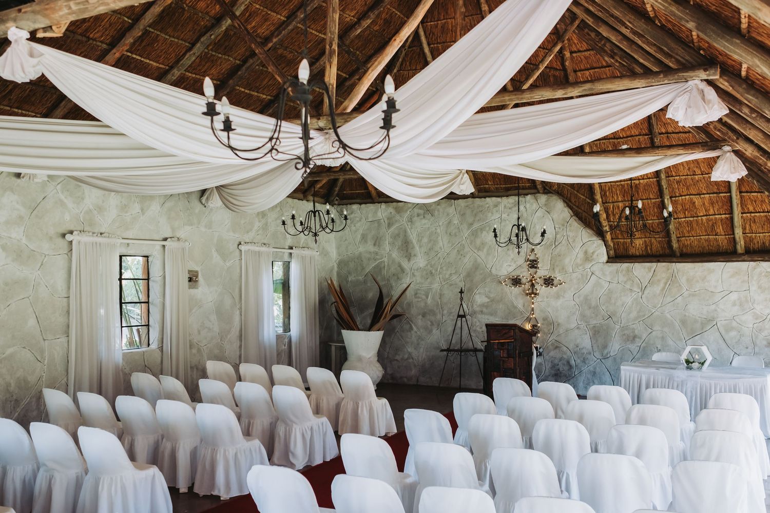Rustic wedding ceremony venue with white draped ceiling, chandelier, and rows of white chair-covered seats.