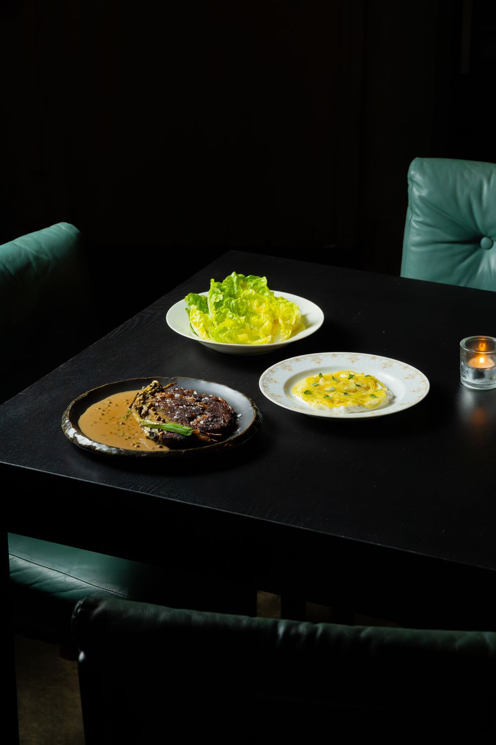 Three plates on a dark table featuring a meat dish with sauce, fresh lettuce, and yellow fruit or vegetables in moody lighting.