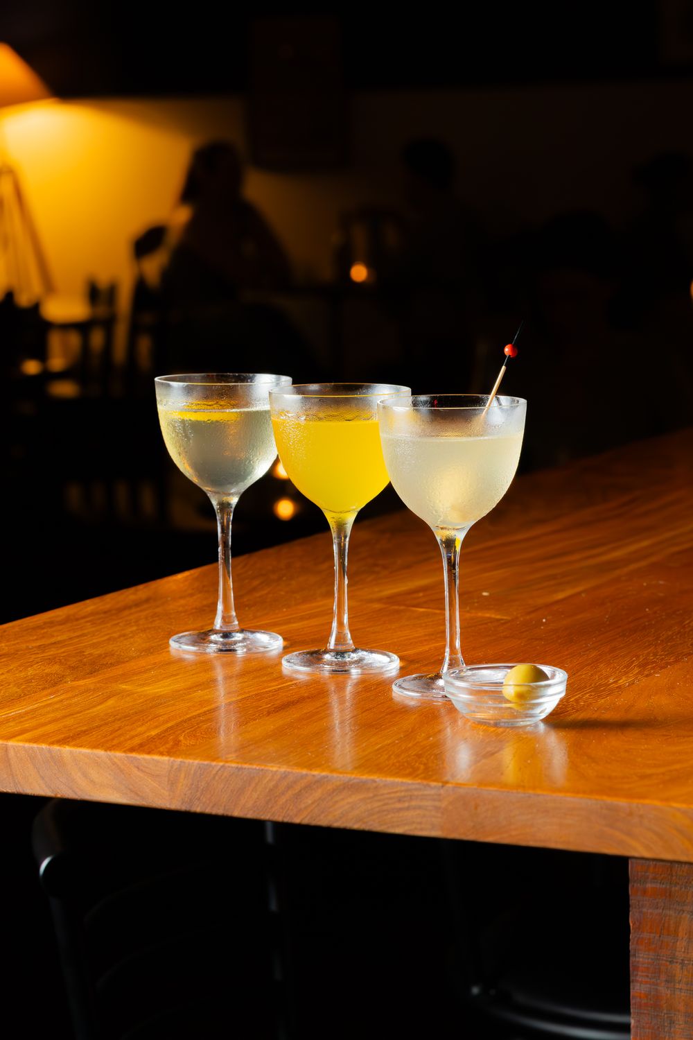 Three elegant cocktail glasses with varying yellow-hued drinks sit on a wooden bar top in moody lighting.