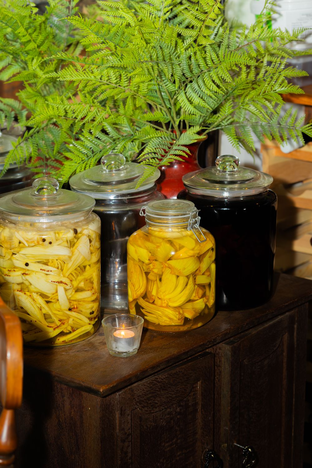 Glass jars filled with preserved yellow and dark fruits sit on a wooden cabinet beneath green fern foliage.