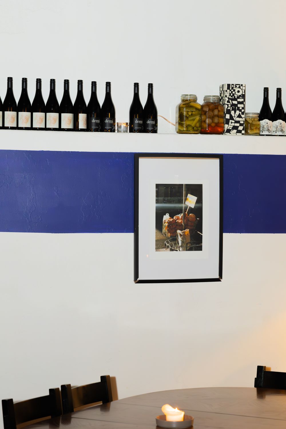 Minimalist dining room with wine bottles displayed on shelf, framed artwork on blue wall stripe, and round table with candle.