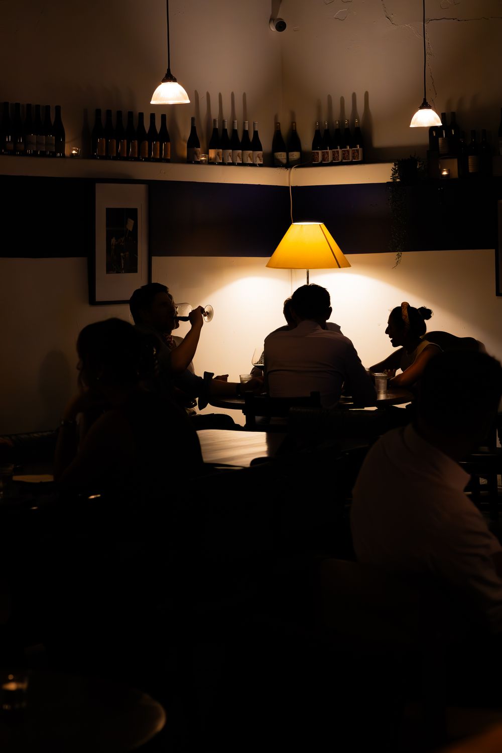Silhouetted figures enjoy drinks at a dimly lit wine bar with bottles on display and warm pendant lighting.