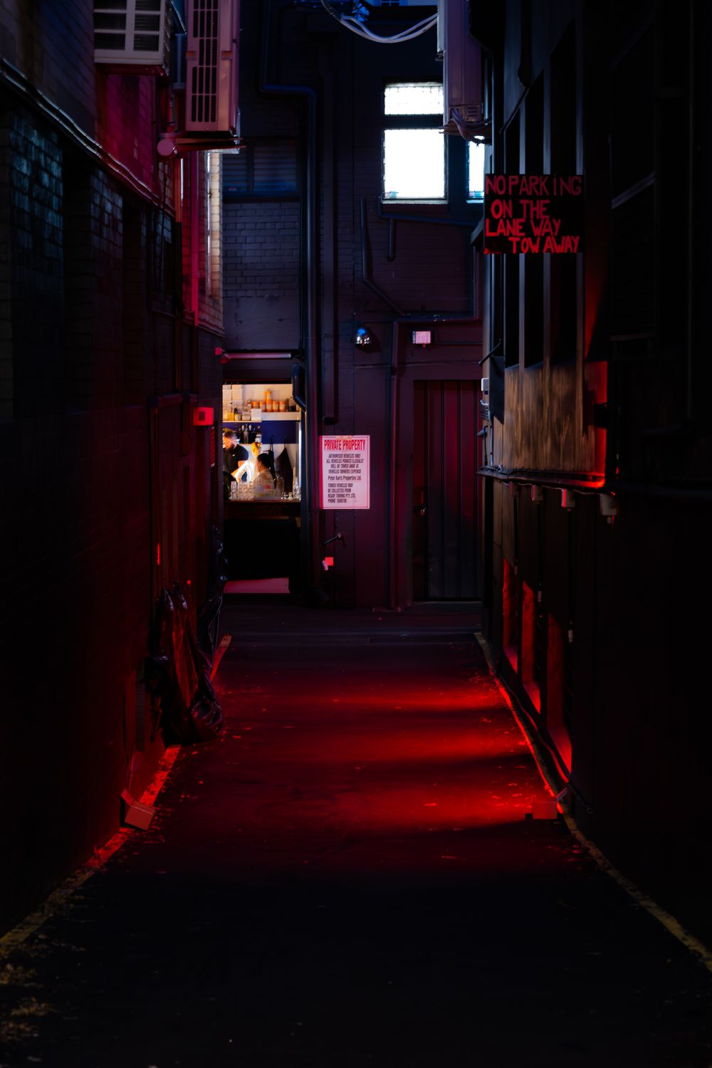 Dark alley illuminated by red neon lights casting an eerie glow on brick walls and windows in urban setting.