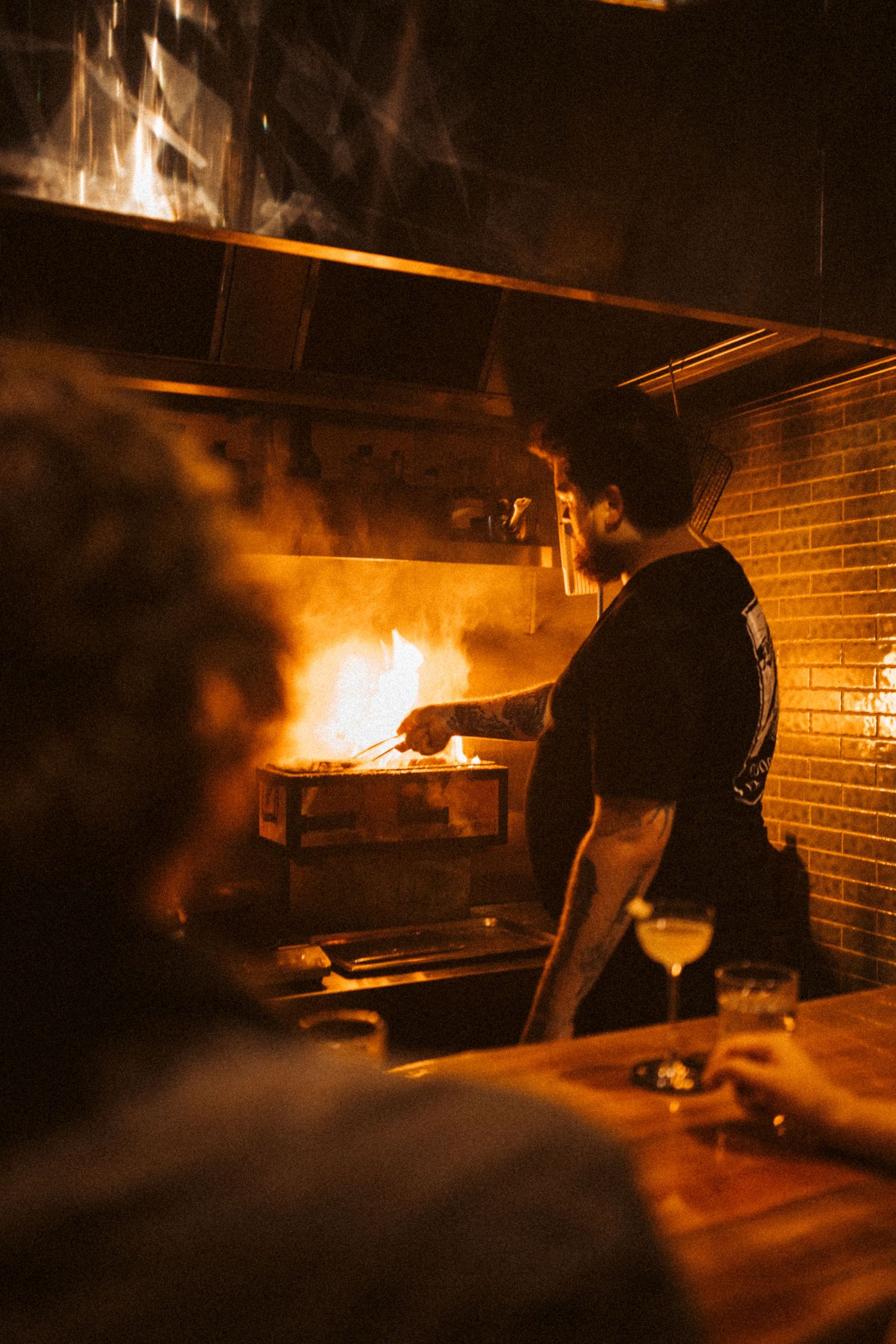 Chef working at a blazing wood-fired brick oven, tending to food with dramatic flames illuminating the kitchen.