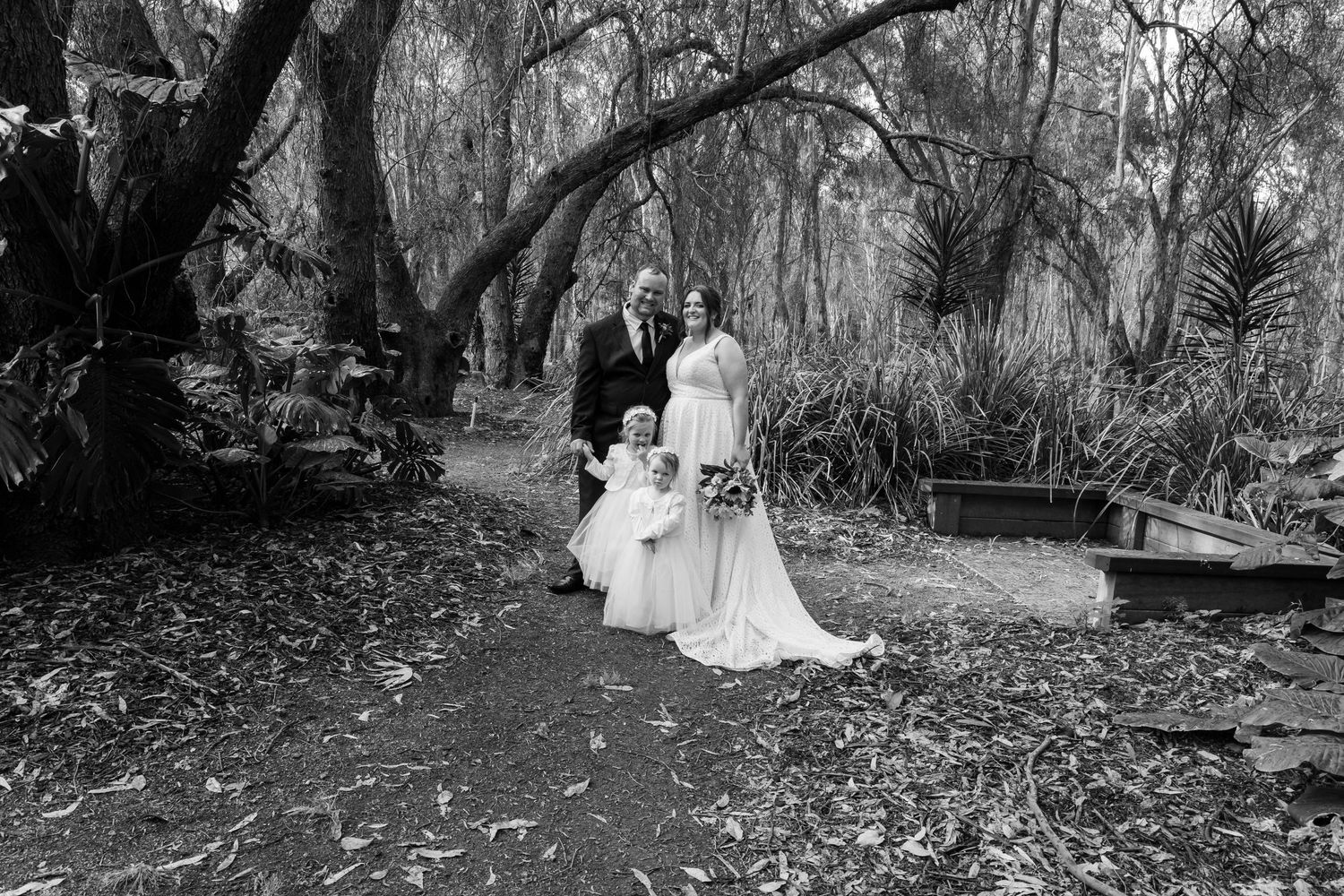 A black and white wedding photograph captured in a lush wooded garden setting.