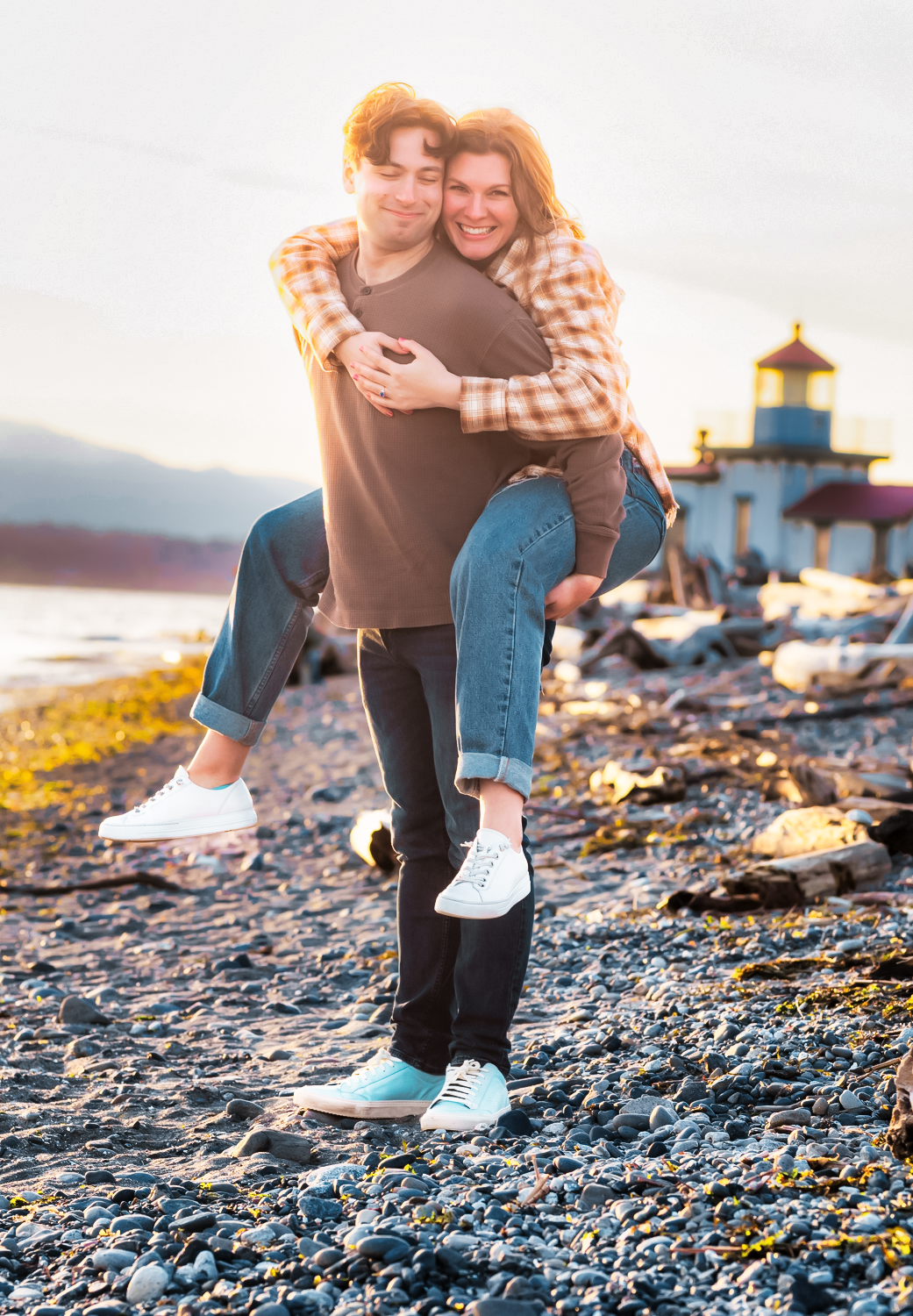 Two figures embrace on a rocky beach shoreline during a dramatic sunset with mountains in the background.