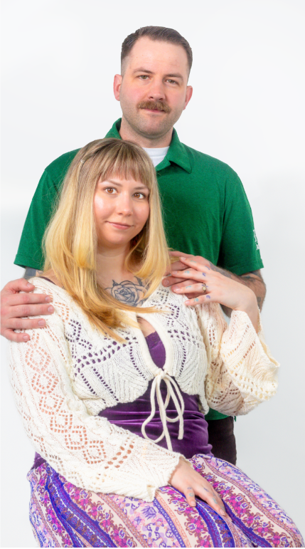 A couple poses for a portrait with the woman wearing a crochet sweater and purple skirt while seated.