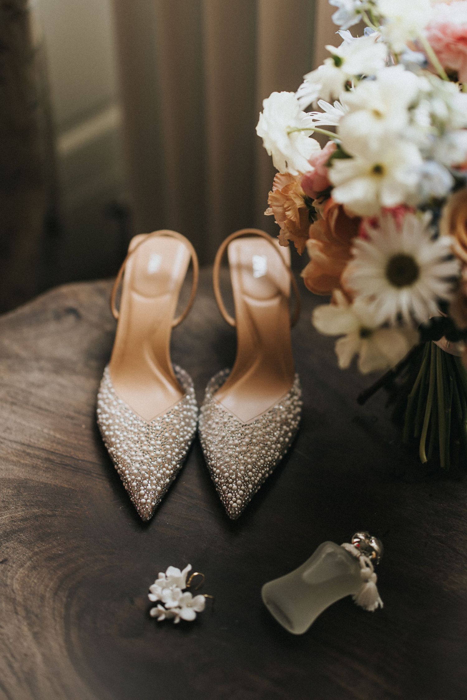 Elegant pearl-studded bridal heels displayed with white daisies and floral arrangements on a dark wooden surface.