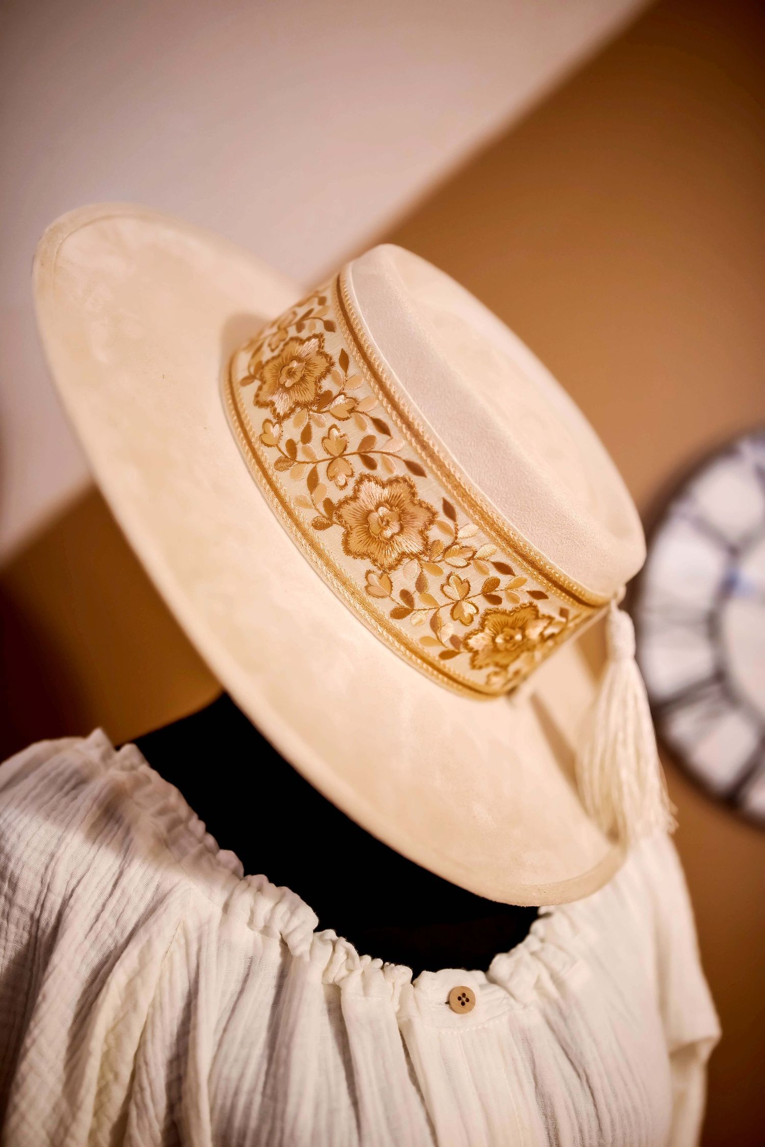 Elegant cream wide-brimmed hat with ornate gold band detailing displayed on a vintage white blouse.