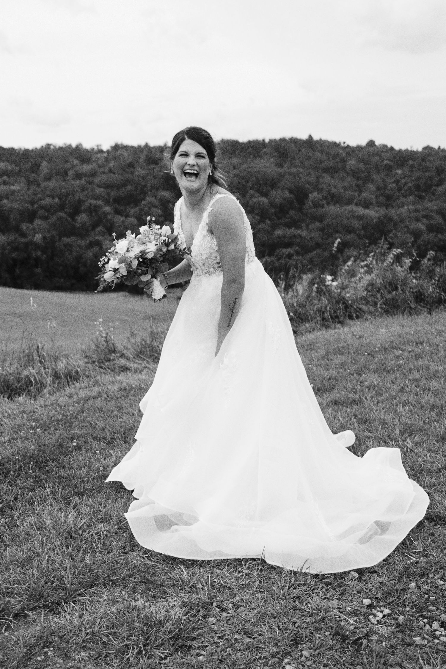 A bride in a flowing white wedding gown twirls joyfully in a grassy field with trees in the background.