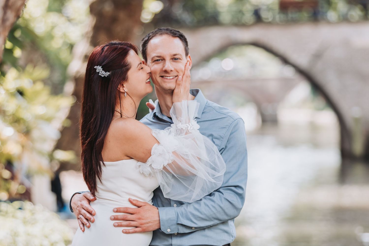 A couple shares a tender moment outdoors in wedding attire with a stone bridge in the background.