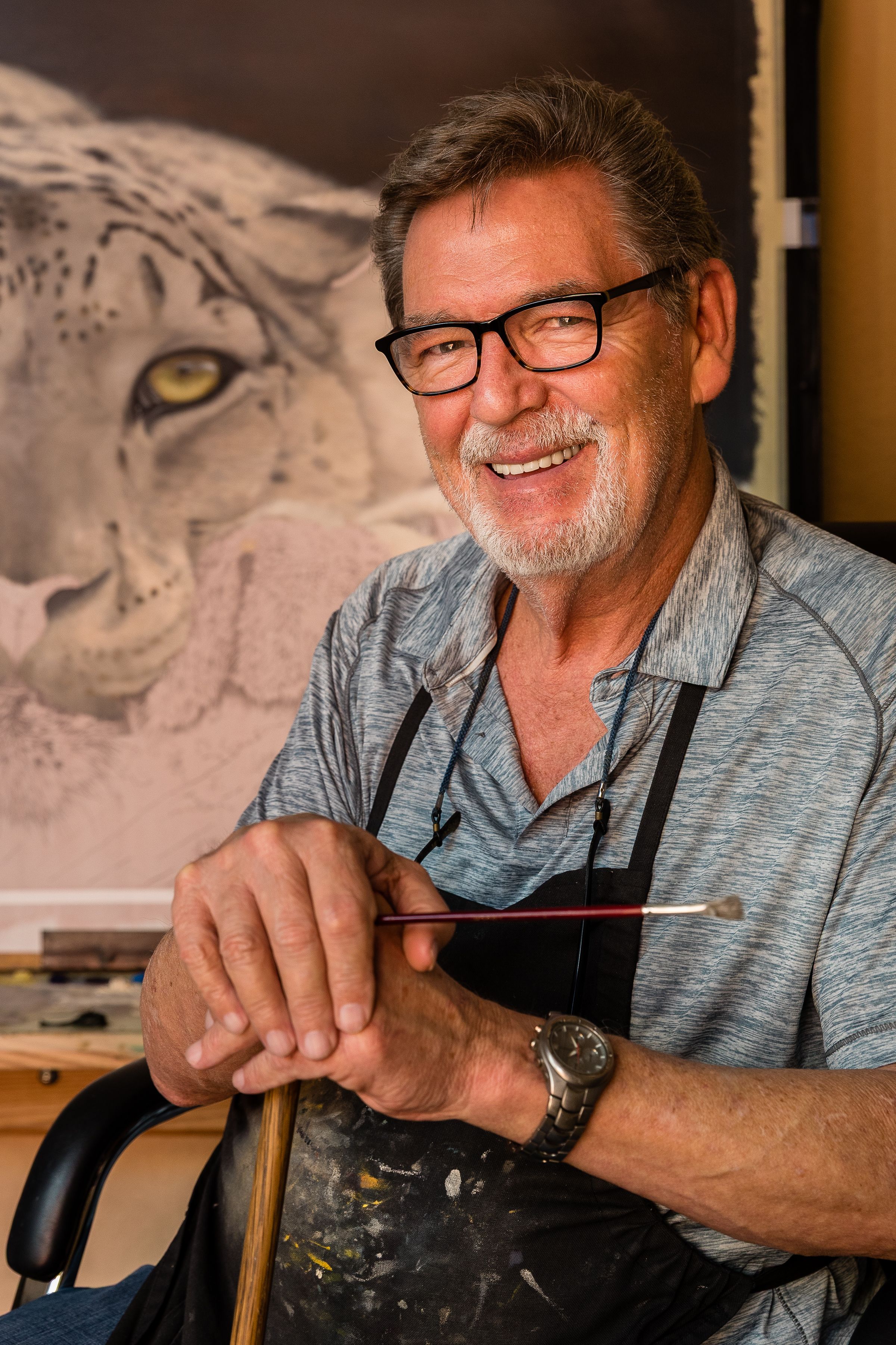 Artist in glasses and an apron smiles while working in their studio with a large wild cat drawing visible in background.