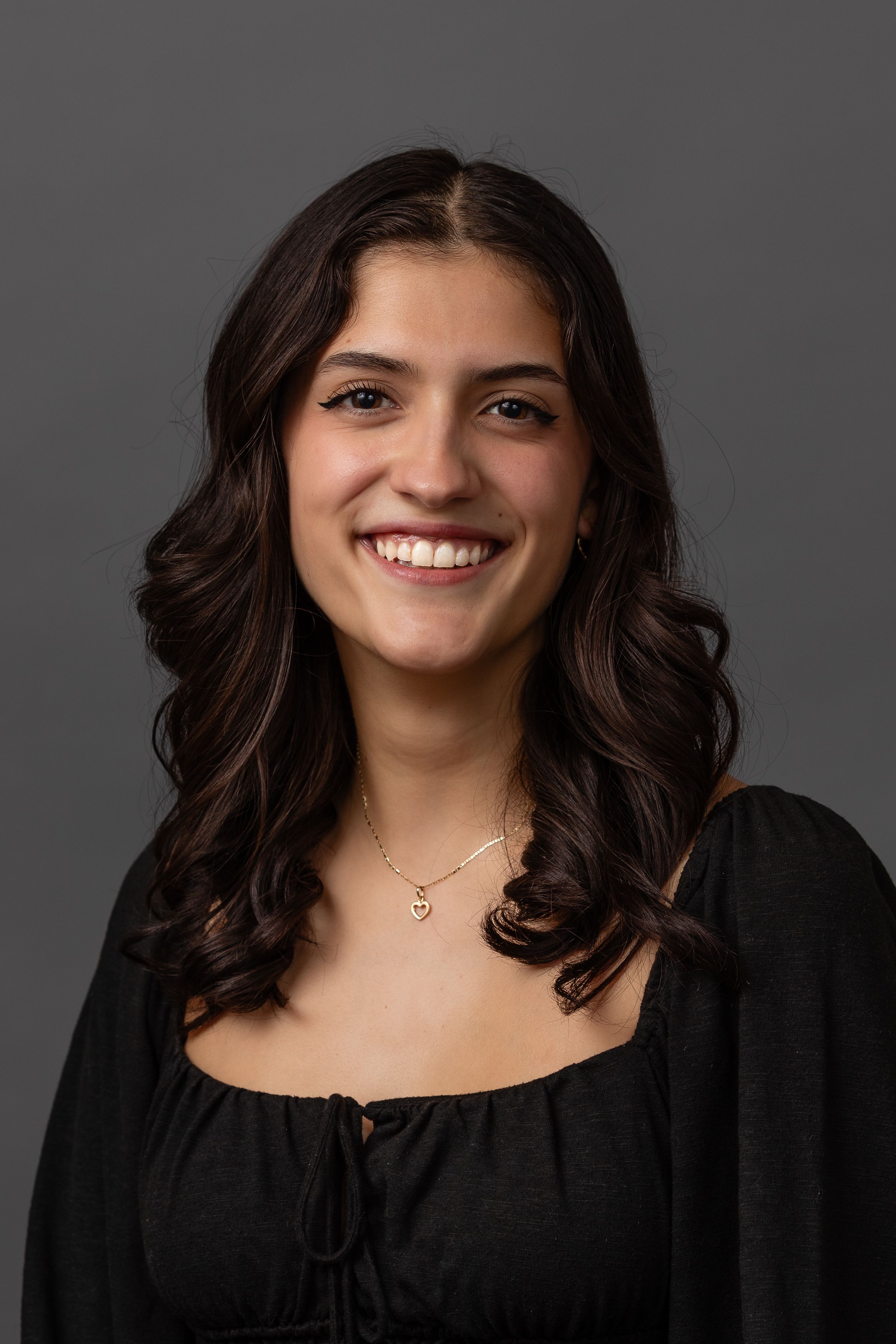 Professional headshot showing someone in a black top against a gray background smiling with dark wavy hair.