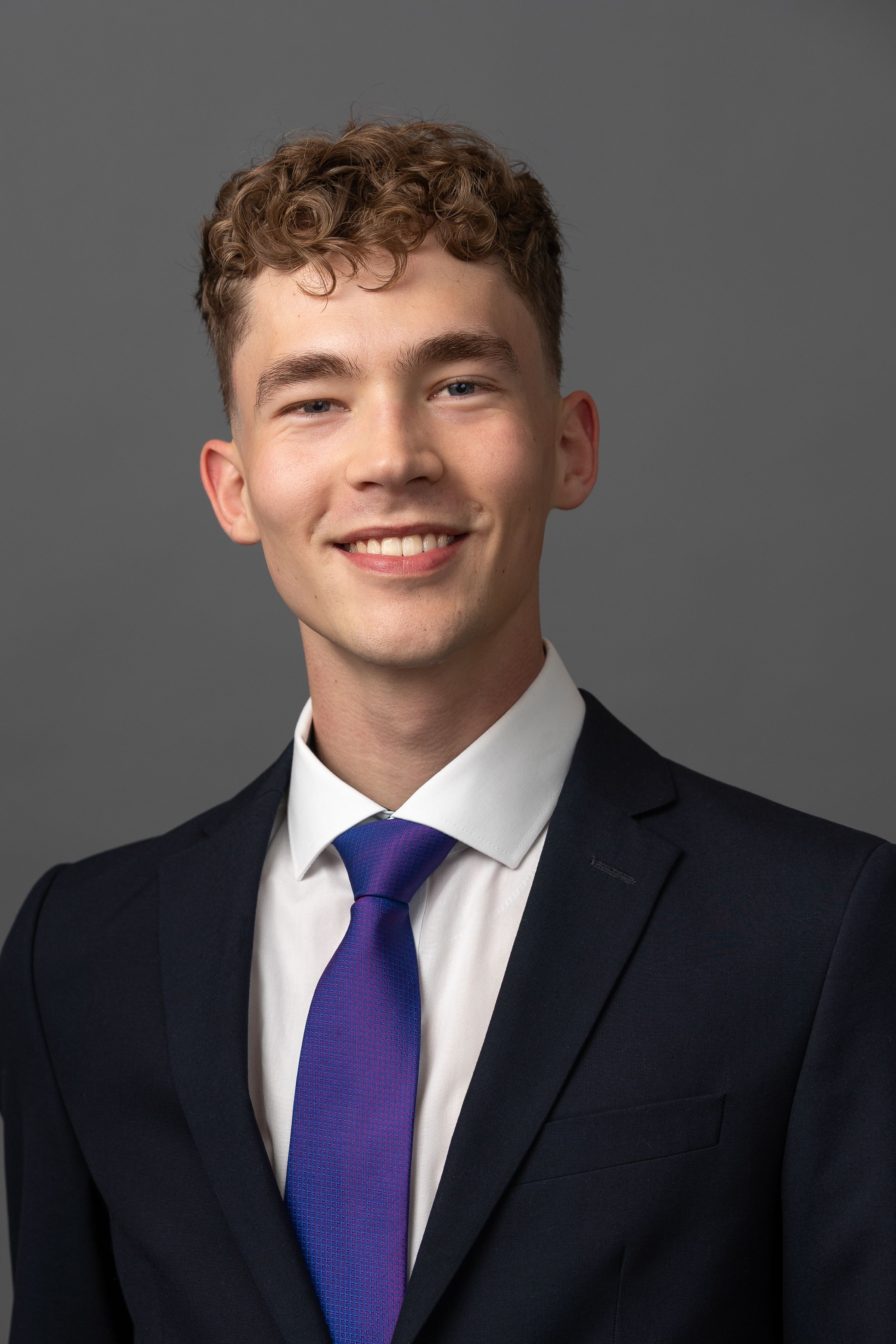 Professional headshot of a person with curly hair wearing a dark suit and purple tie against a grey background.
